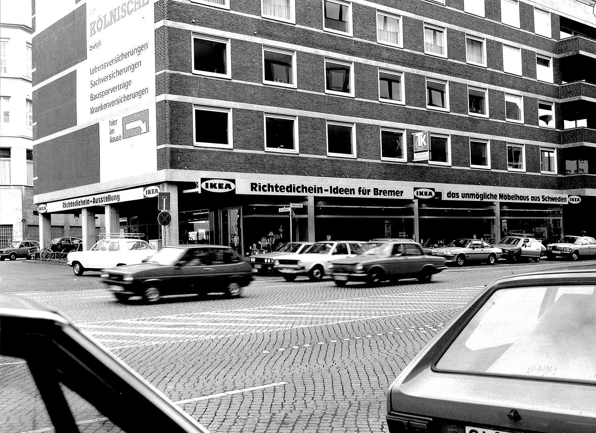 Black and white street scene with an IKEA store, KÖLNISCH signs, and cars on a cobblestone road.