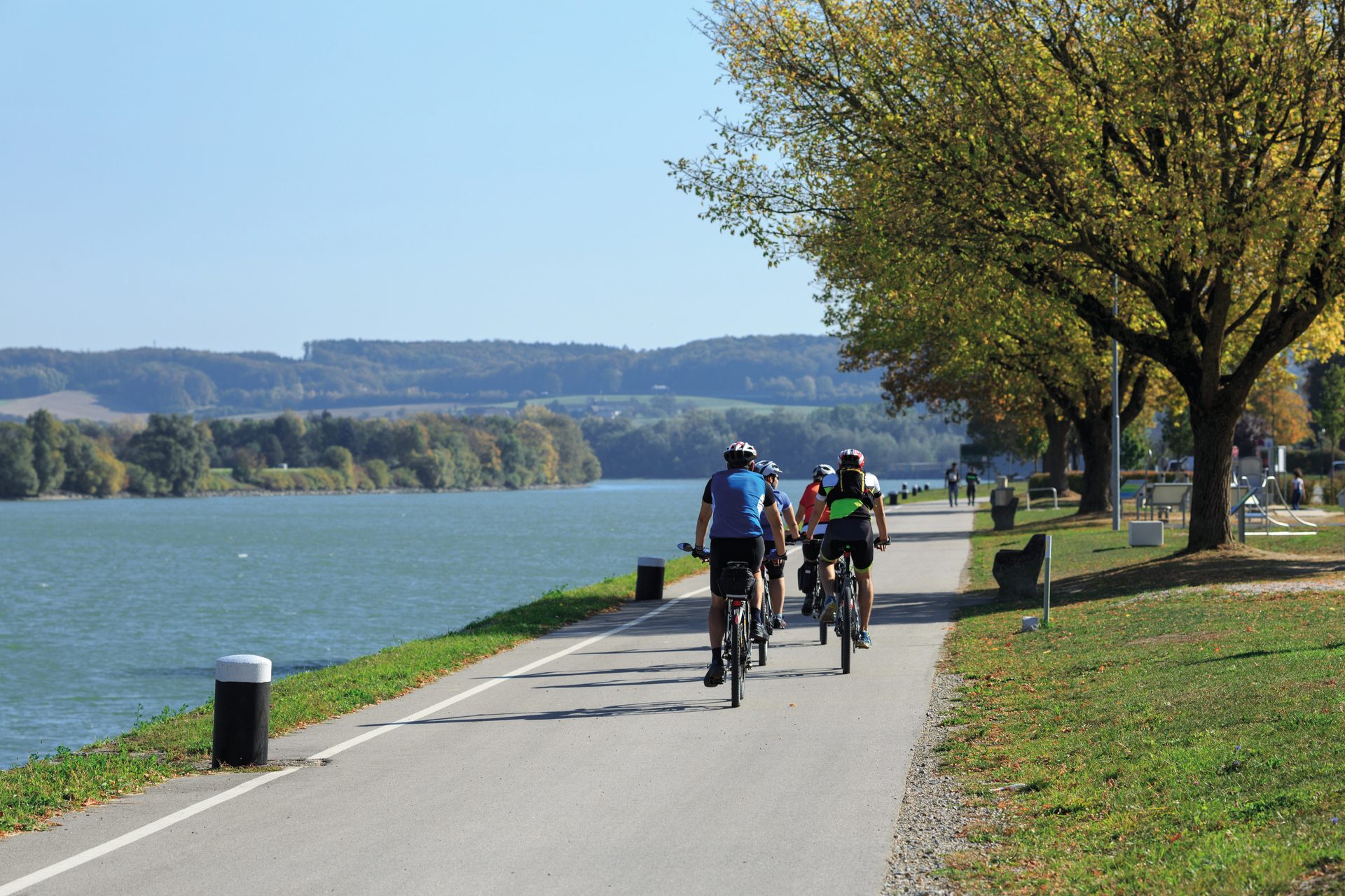 Cyclists on a paved path next to a wide river, with autumn trees and distant hills under a clear sky.