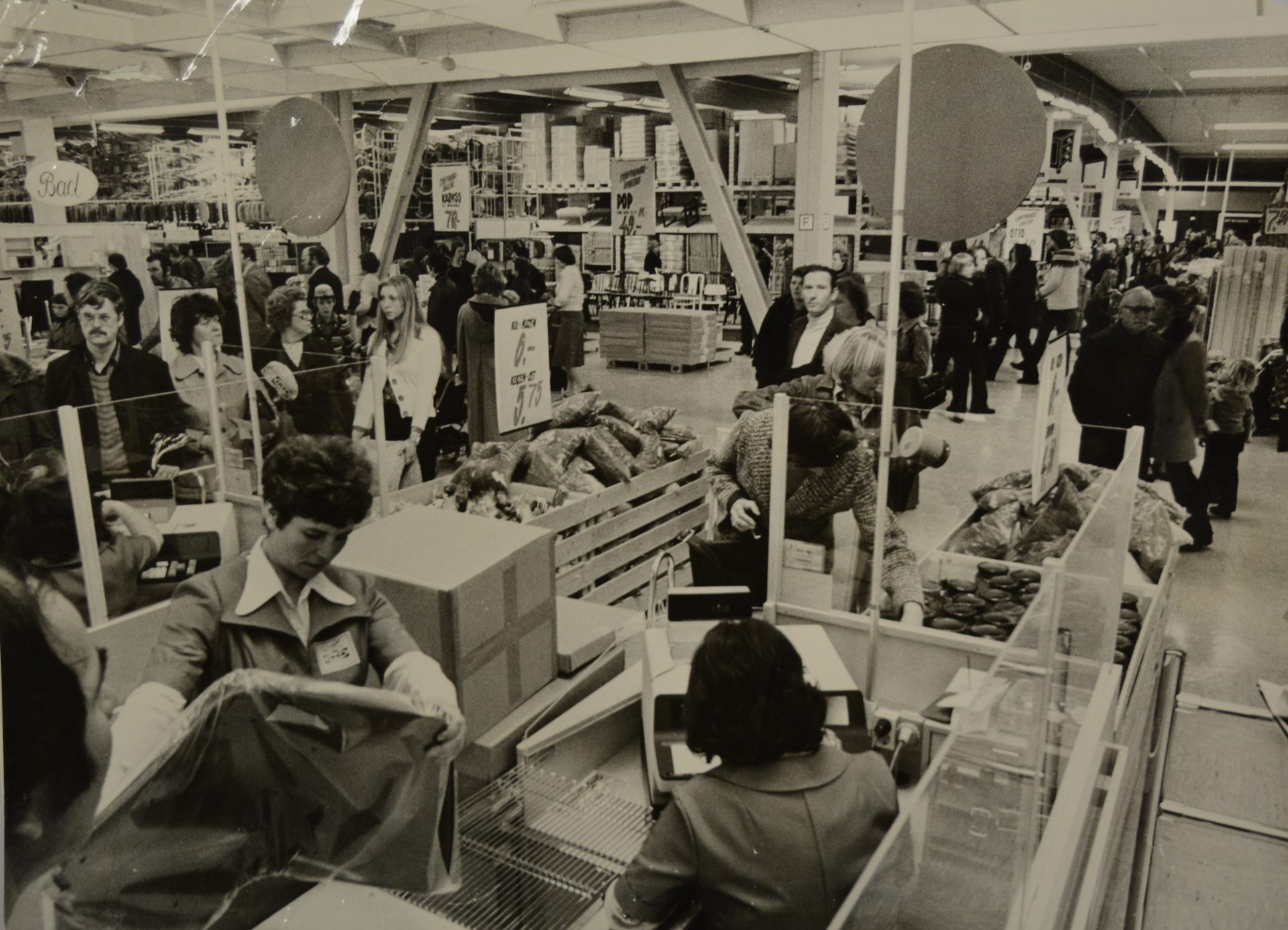 Black and white photo of a busy supermarket checkout area with customers and cashiers.