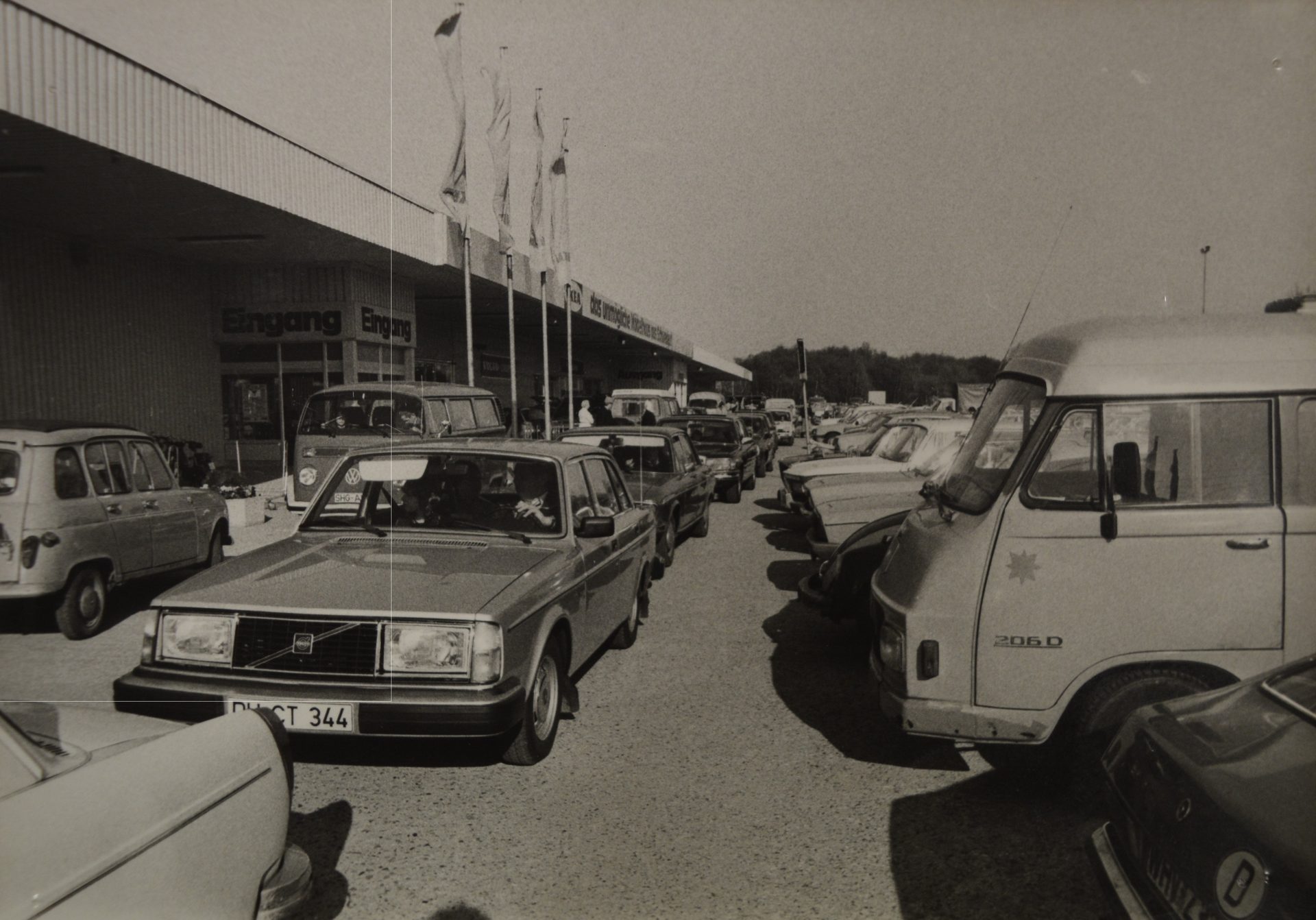 Black and white photo of a crowded parking lot with vintage cars outside a building with "Eingang" signs.