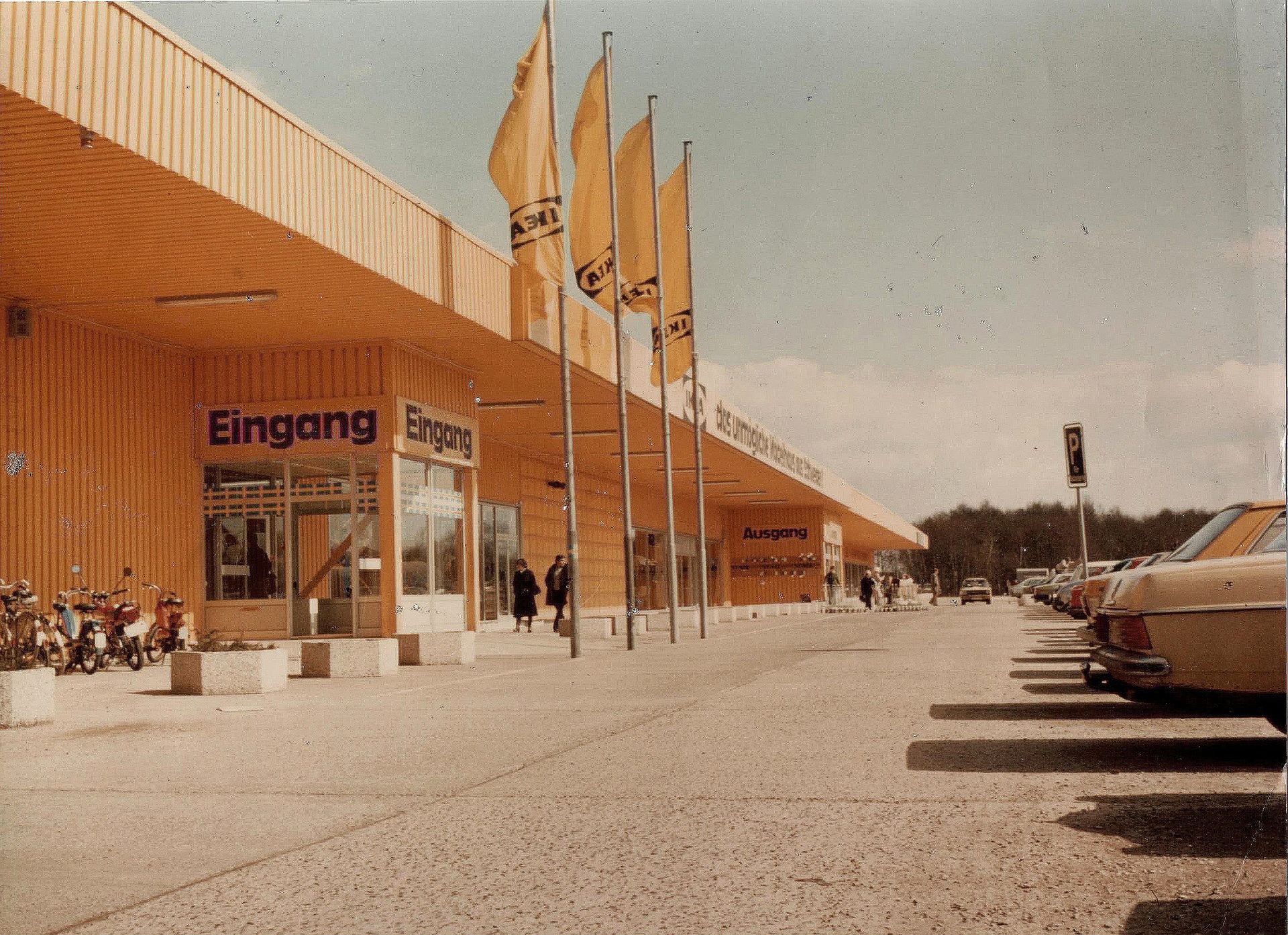 A vintage photo of a yellow IKEA store exterior with an "Eingang" sign, flags, and parked cars.