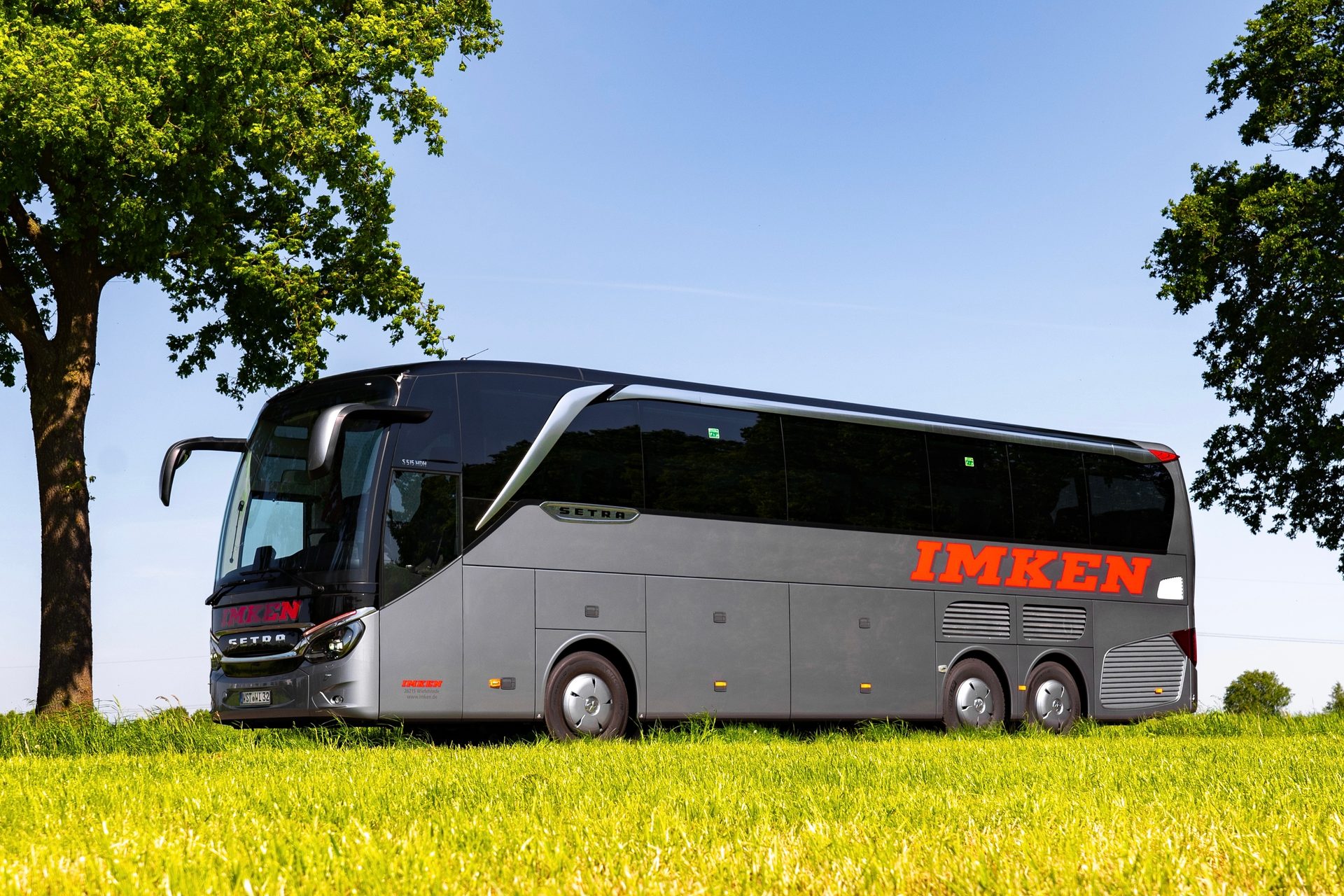 A gray and black Setra Imken coach bus parked in a green field under a blue sky, framed by trees.