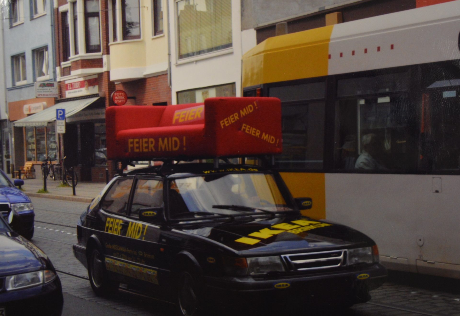 A black car with a large red sofa on its roof, advertising "FEIER MID!", drives past a tram and buildings.