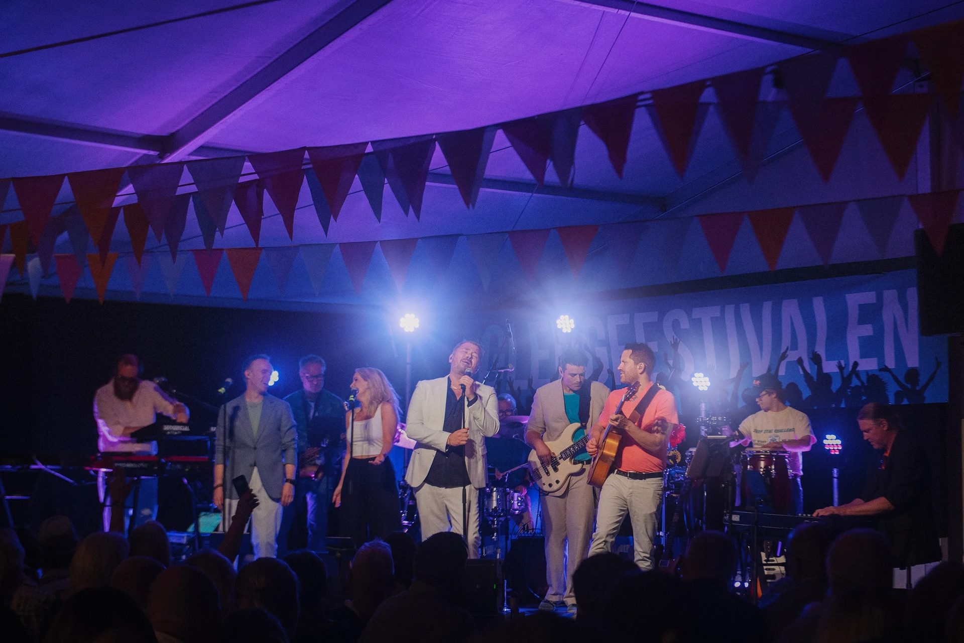 A band performs live on stage under purple lights, featuring multiple singers and musicians playing instruments.