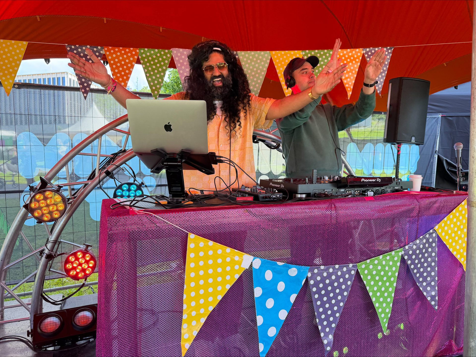 Two lively DJs perform at an outdoor event under a red tent, surrounded by colorful bunting.