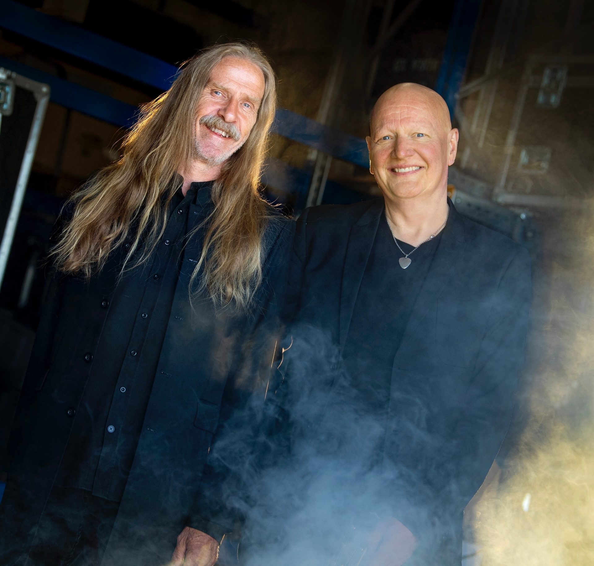 Two smiling men, one with long hair, one bald, in dark clothes with smoke.