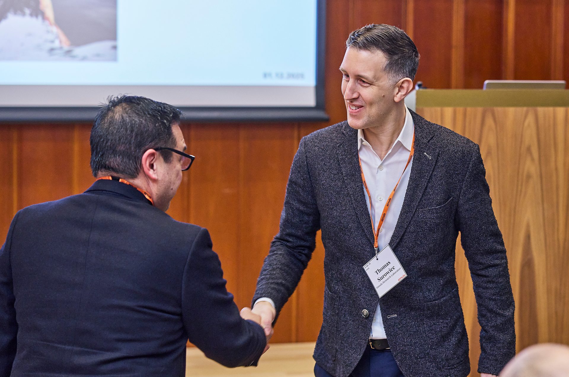Two men shaking hands, one smiling, with an ID tag showing 'Thomas Surowiec'.