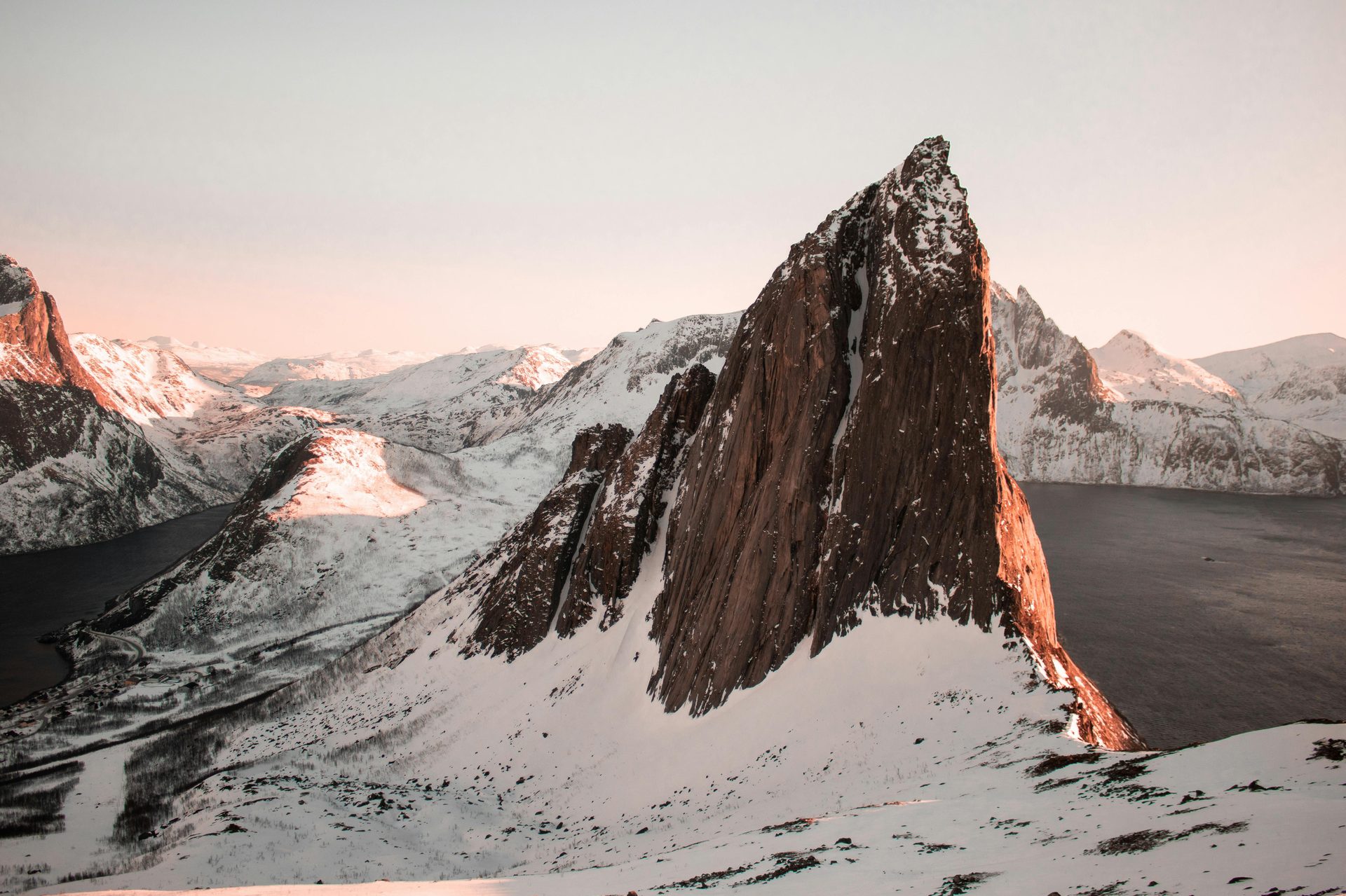 Dramatic snowy mountains with a jagged peak bathed in warm light, overlooking a fjord.