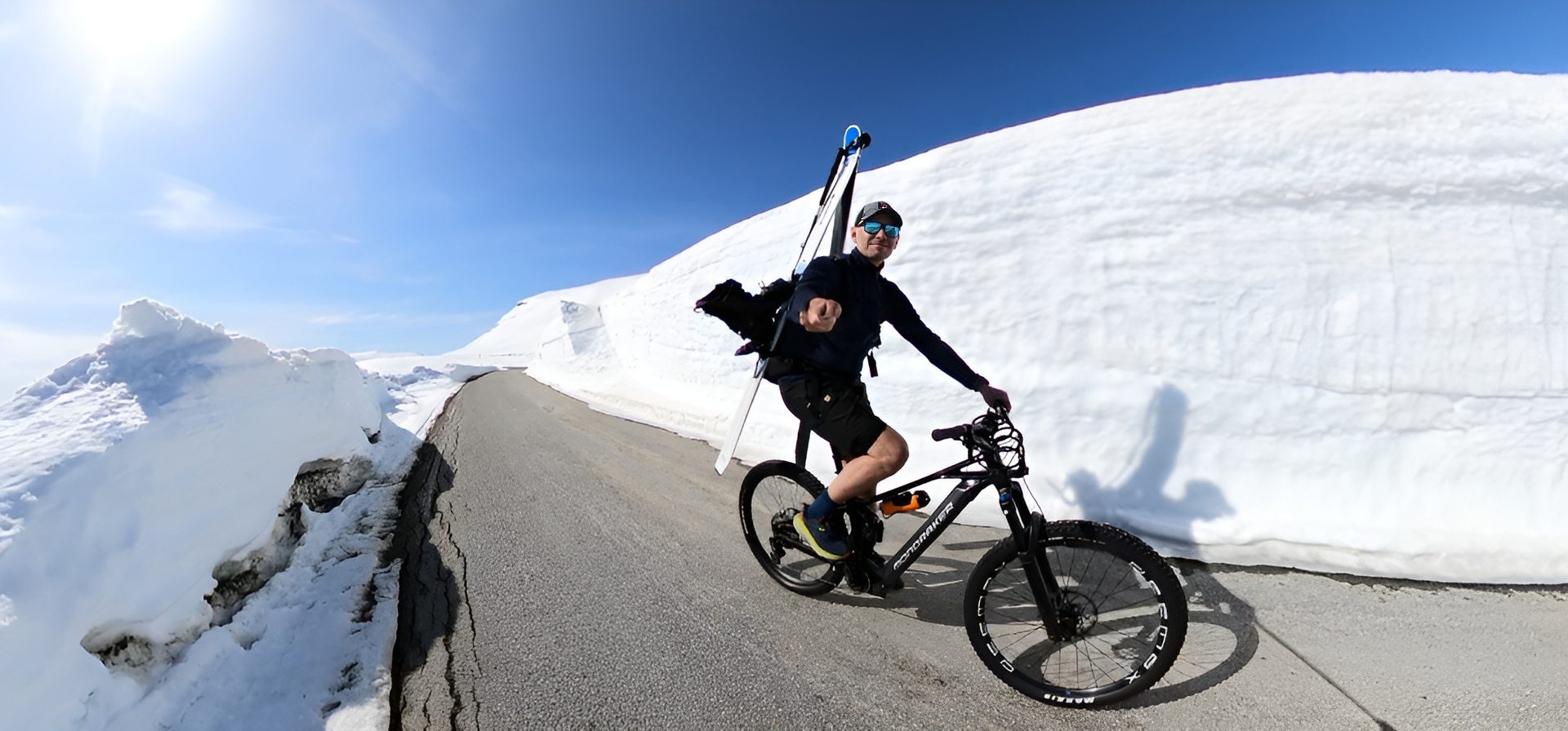 Man on bike with skis on back, riding on a road between massive snow walls under blue sky.