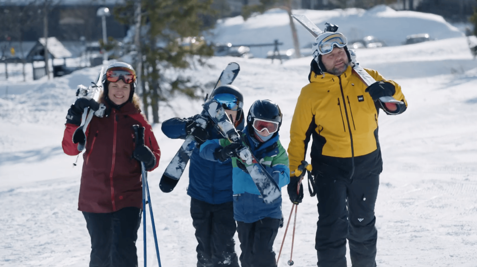 Smiling family of four in ski gear, carrying skis and poles, on a snowy slope.