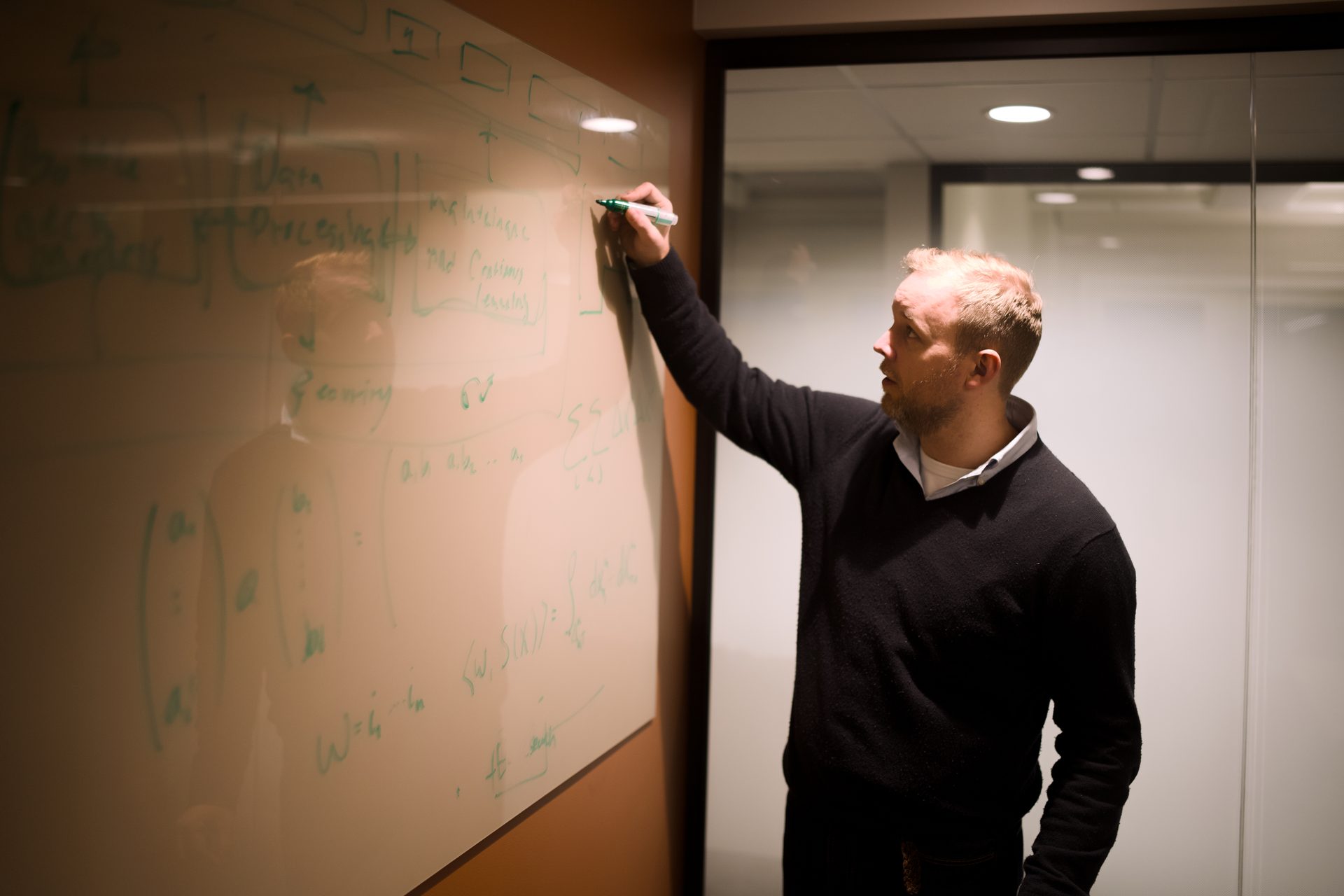 Man in an office writes complex diagrams and equations on a whiteboard with a green marker.