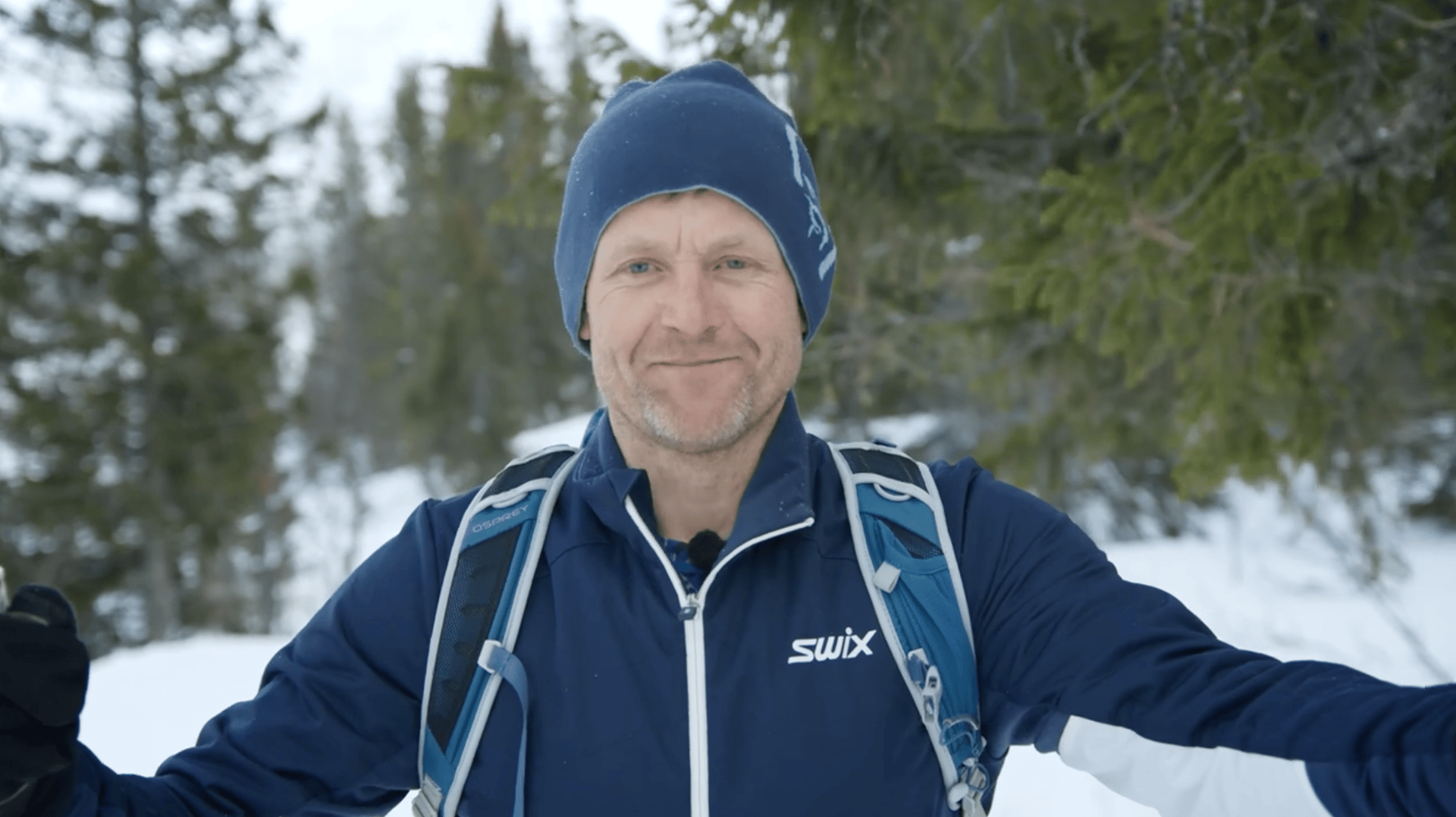 Smiling man in blue winter gear and backpack in a snowy forest.