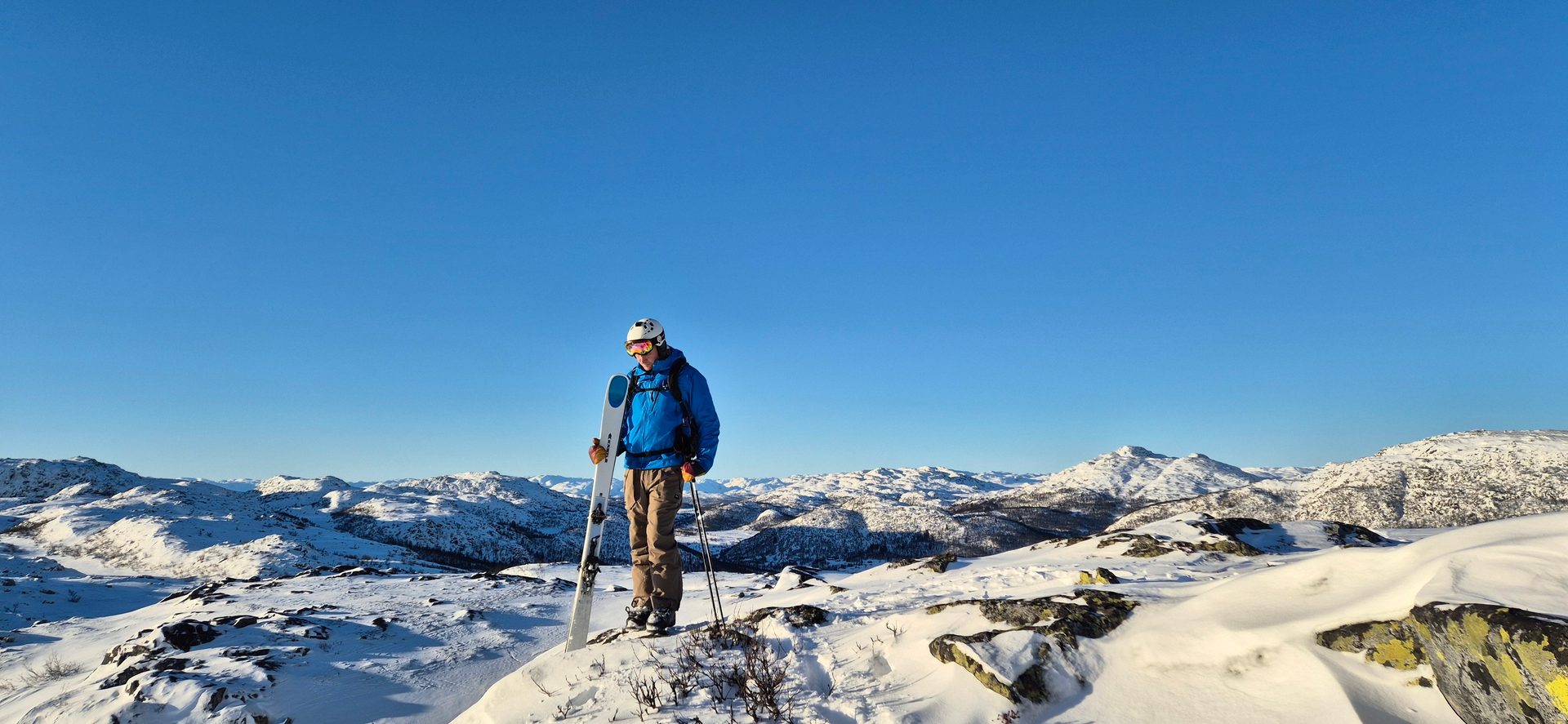 Skier in blue jacket, helmet, skis, and poles on a snowy mountain overlooking a winter landscape, blue sky.