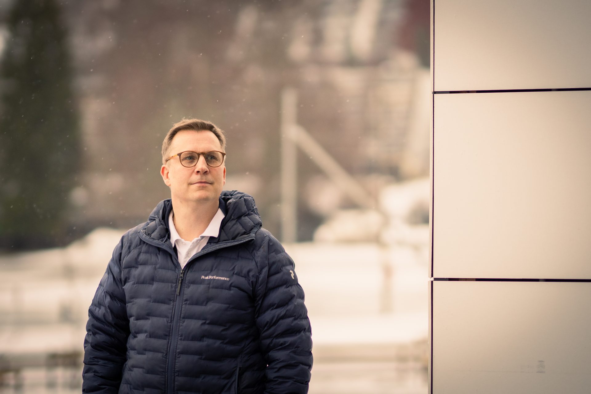 Man in glasses and dark blue puffer jacket, looking right, with blurred snowy background.