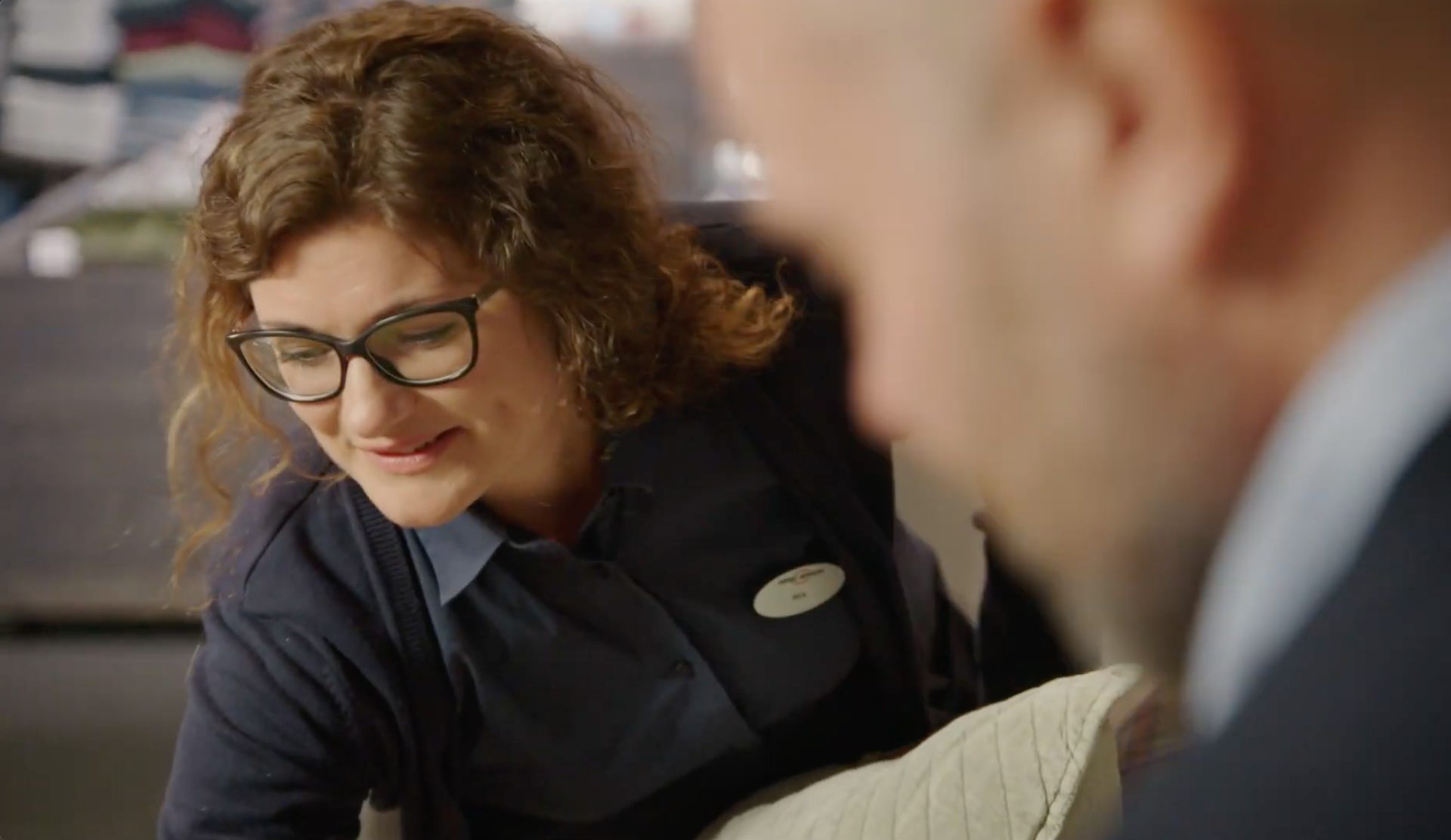 A smiling woman with glasses and curly hair looks down, with a blurry man in the foreground.
