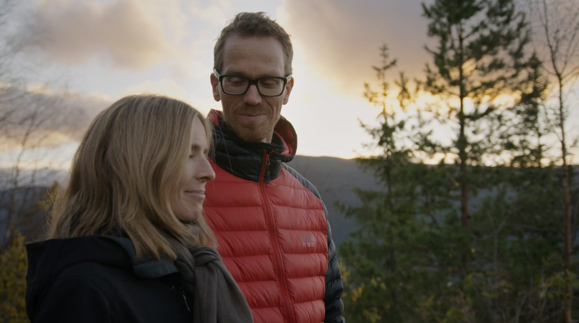 Two people, a man in a red jacket and a woman, standing outdoors at sunset with trees and hills.