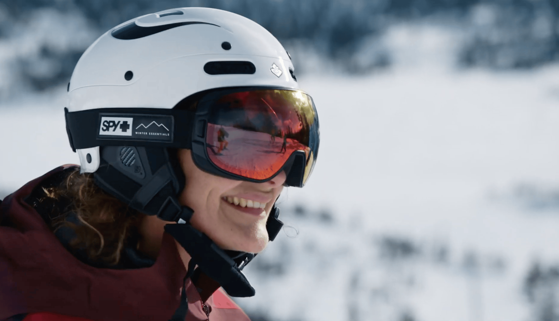 Smiling skier in white helmet and reflective SPY+ goggles against a snowy mountain.