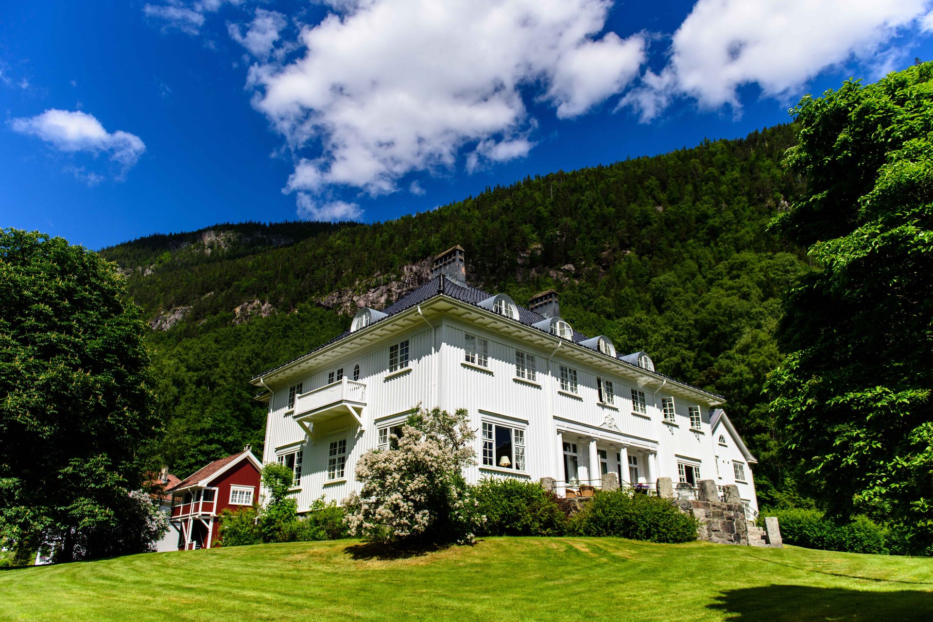 A large white house on a green lawn, backed by a lush forested mountain under a blue sky with clouds.