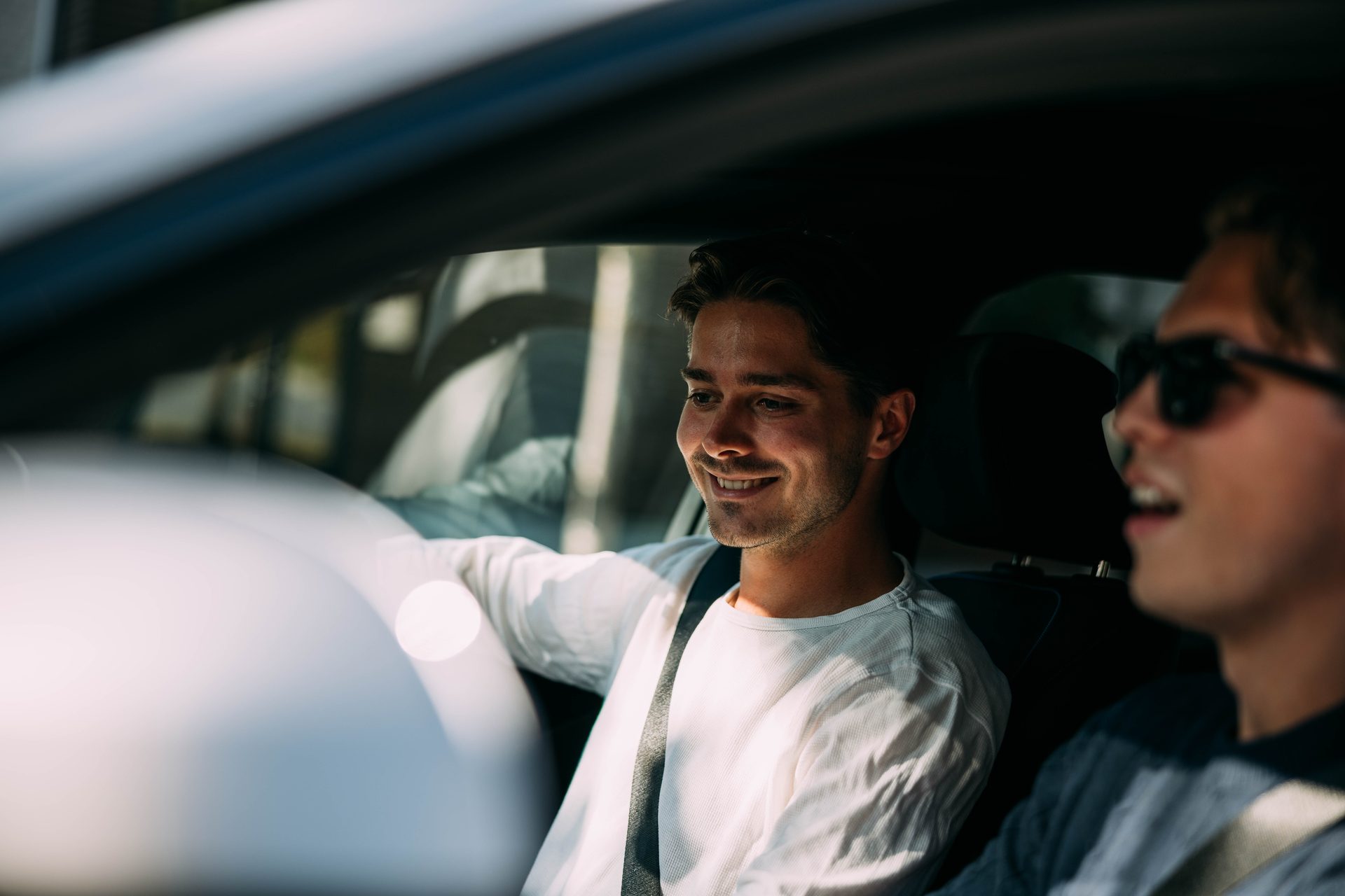 Two men in a car, the driver smiling while a passenger talks, enjoying a ride on a sunny day.