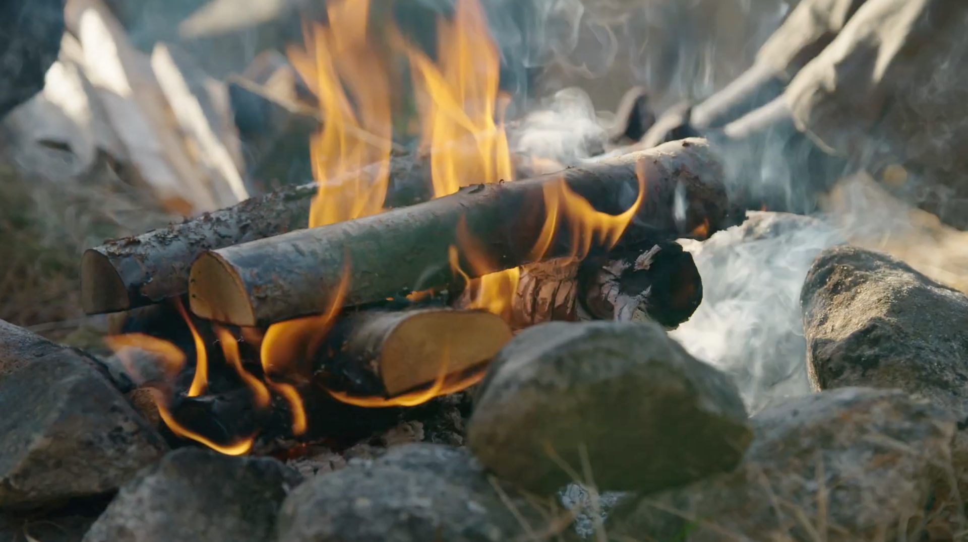 Close-up of a campfire with bright flames, logs, smoke, and surrounding rocks.