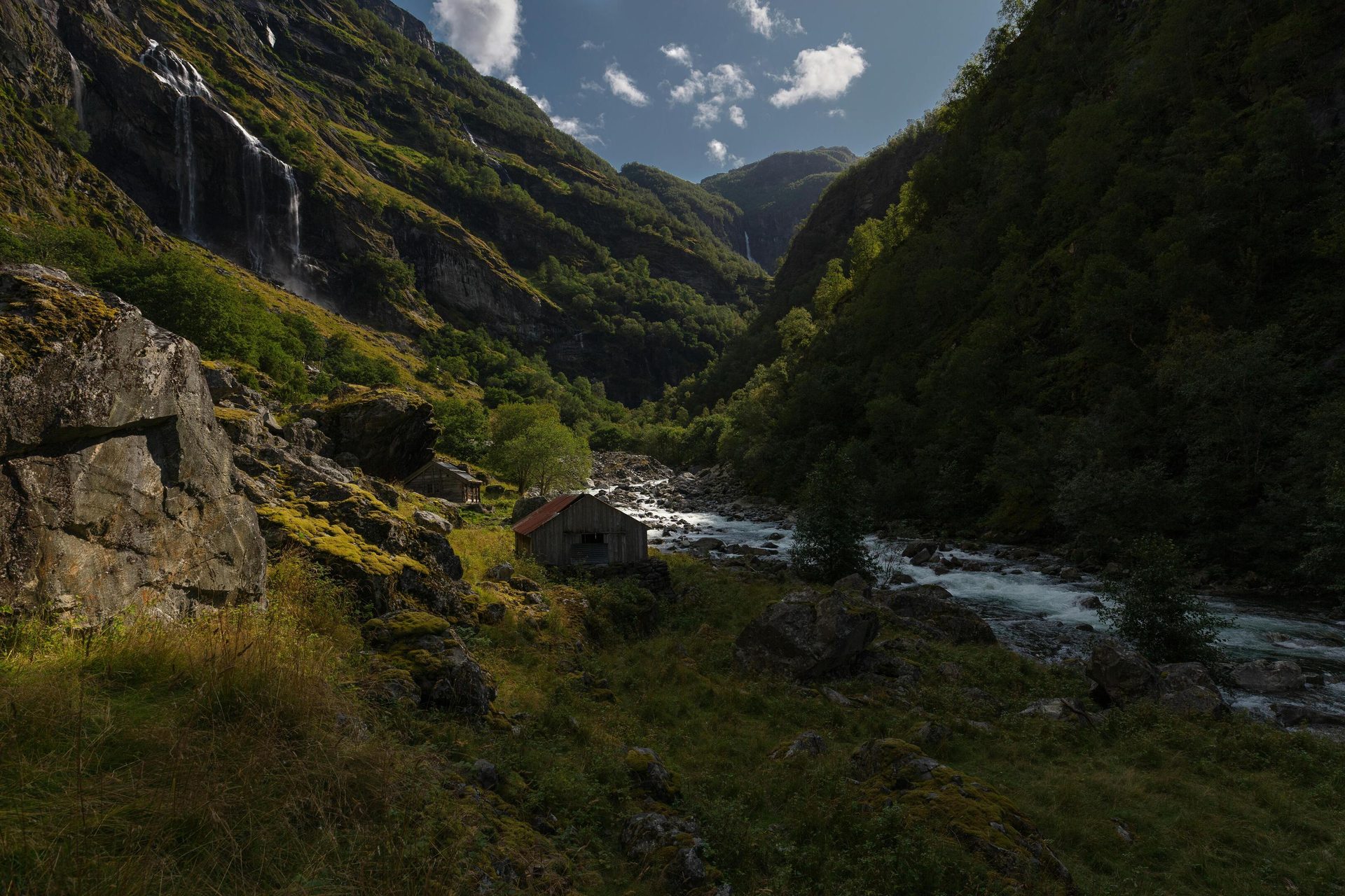 Steep green valley with waterfalls, a river, and rustic wooden cabins.