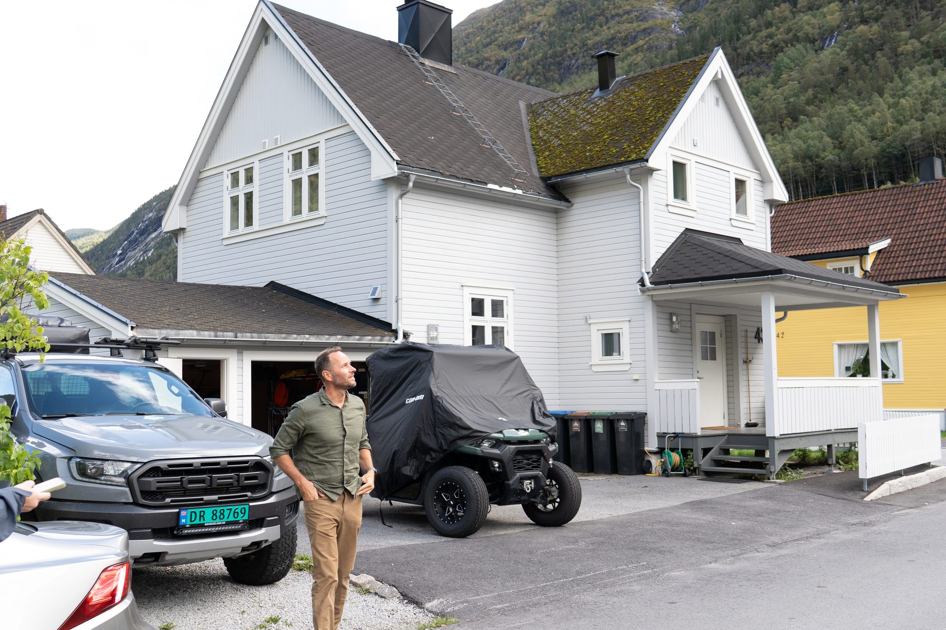 Man standing near a gray pickup truck and a covered ATV in front of a light grey house, mountains behind.