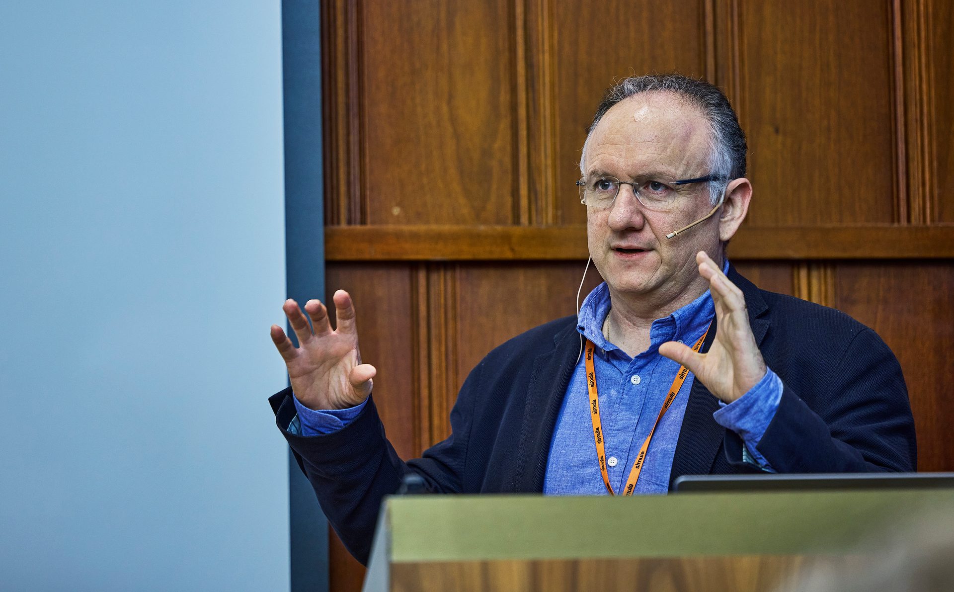 Man in blue shirt and glasses speaking, gesturing with both hands.