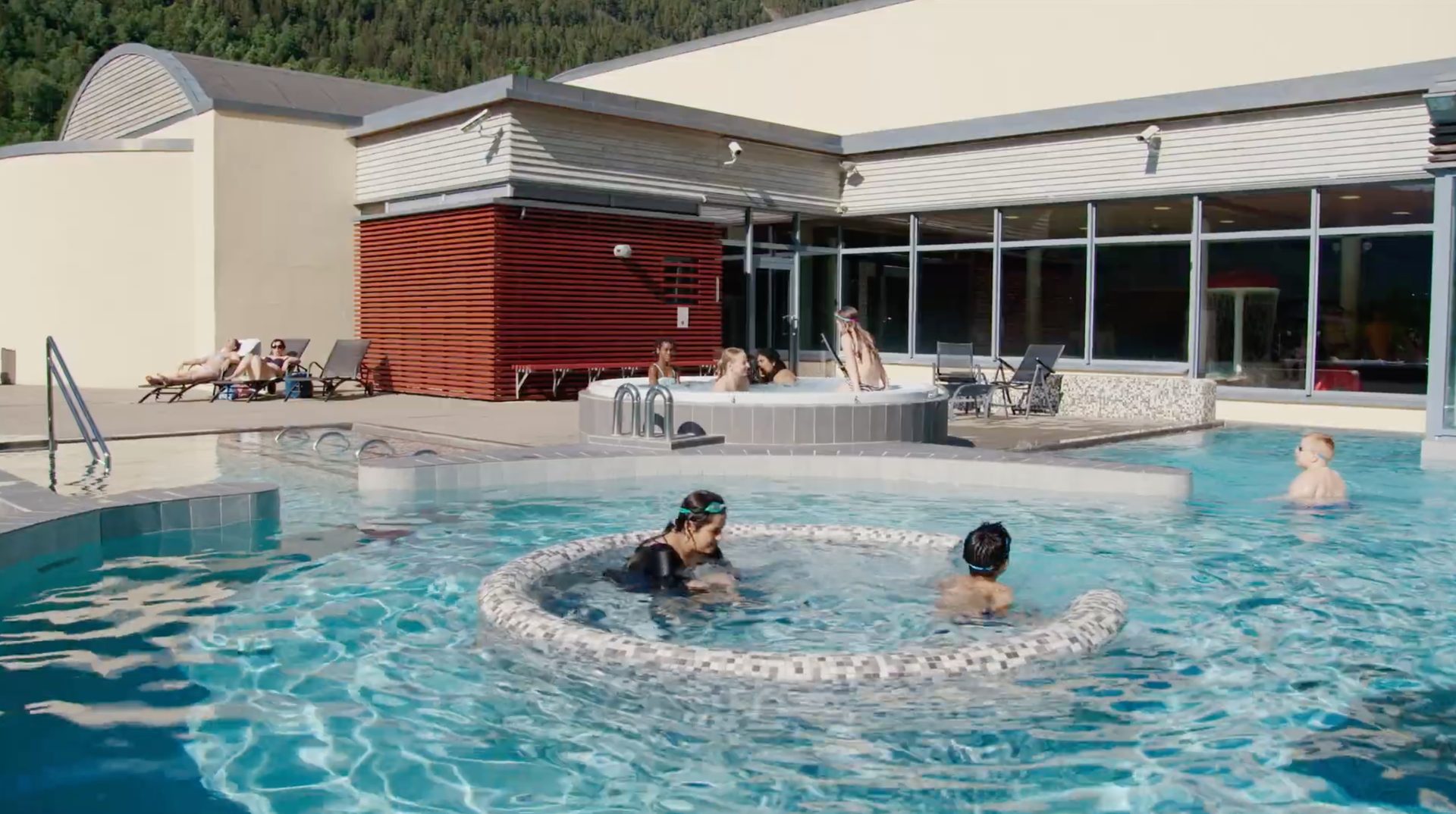 Outdoor pool complex: people in pools, hot tub, sunbathing. Modern building, mountain backdrop.