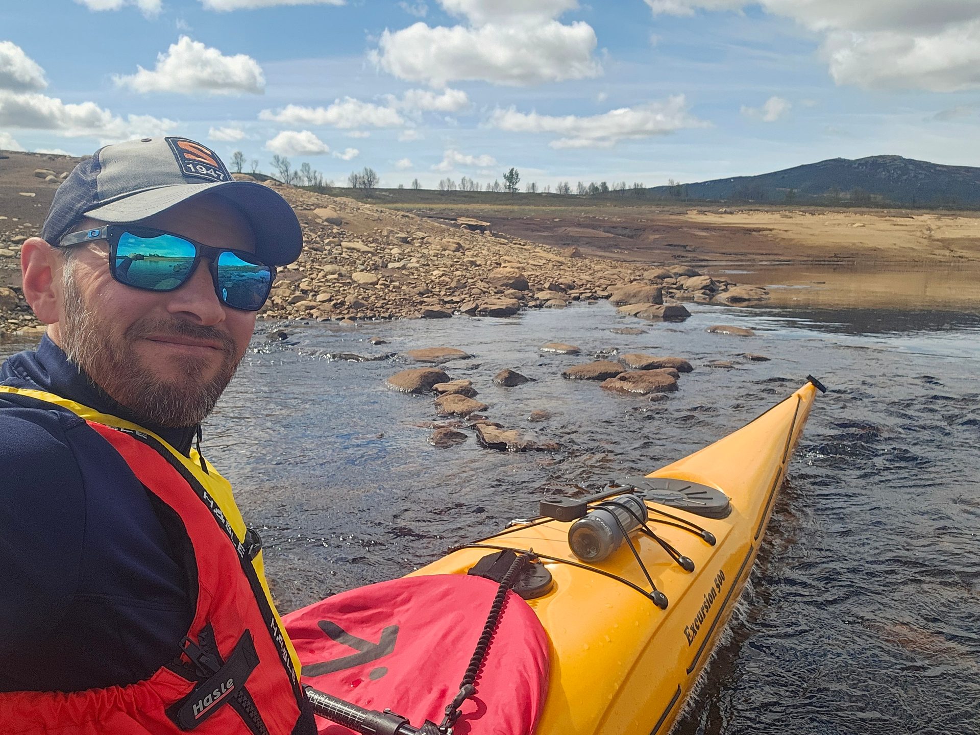 Man in a yellow kayak on rocky water with a life vest, sunglasses, and hat, under a cloudy sky.