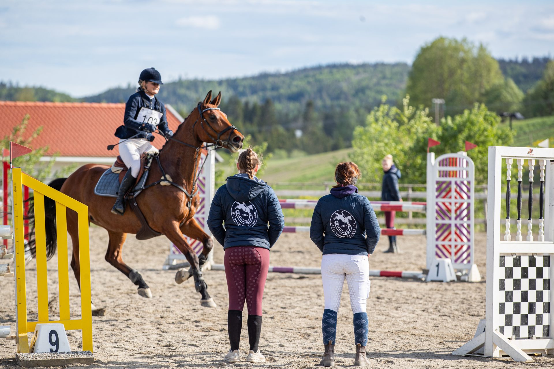 A horse and rider jump an obstacle, while two people watch from behind at an equestrian event.