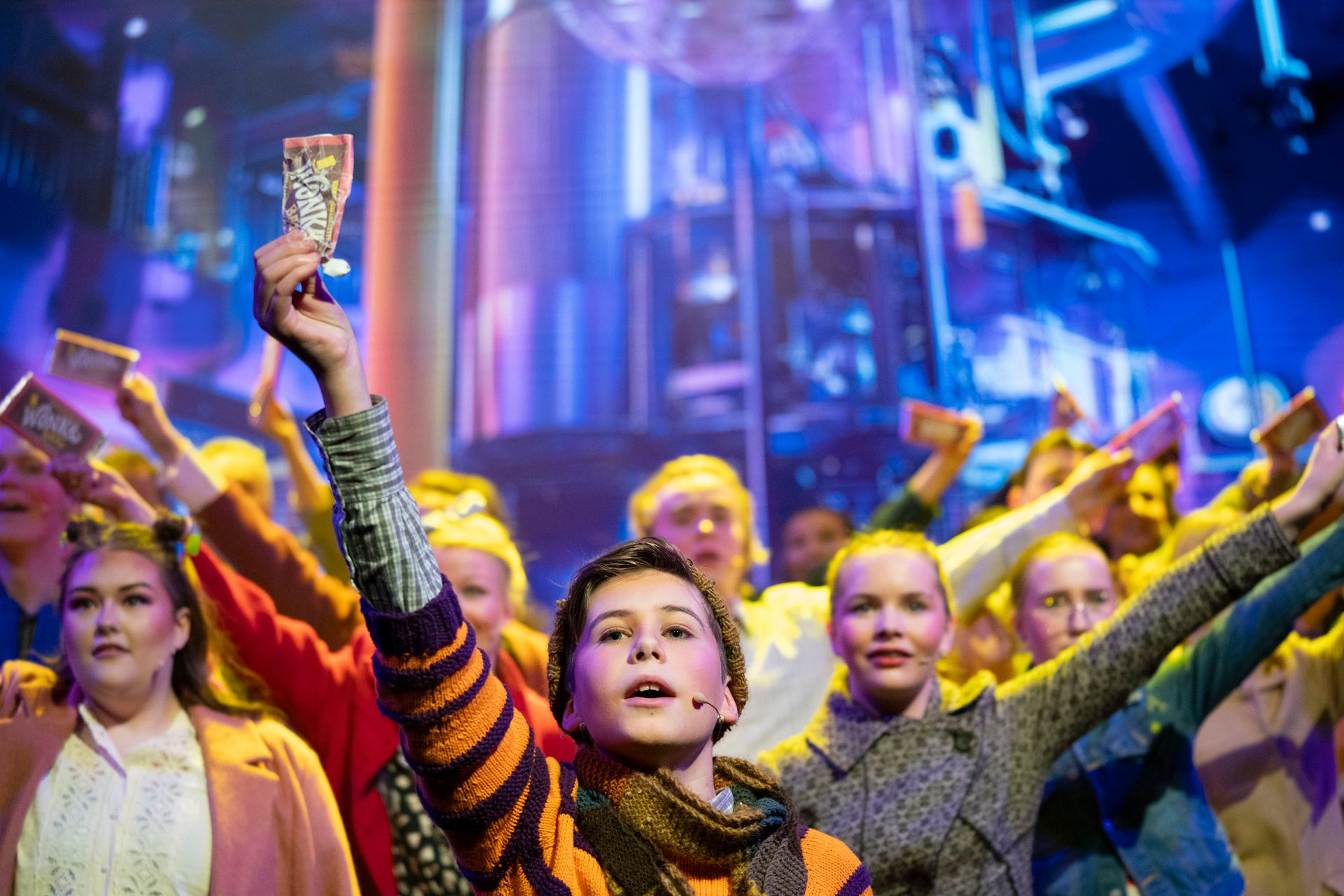 Young person on stage holding a Wonka bar wrapper, surrounded by other performers holding wrappers.
