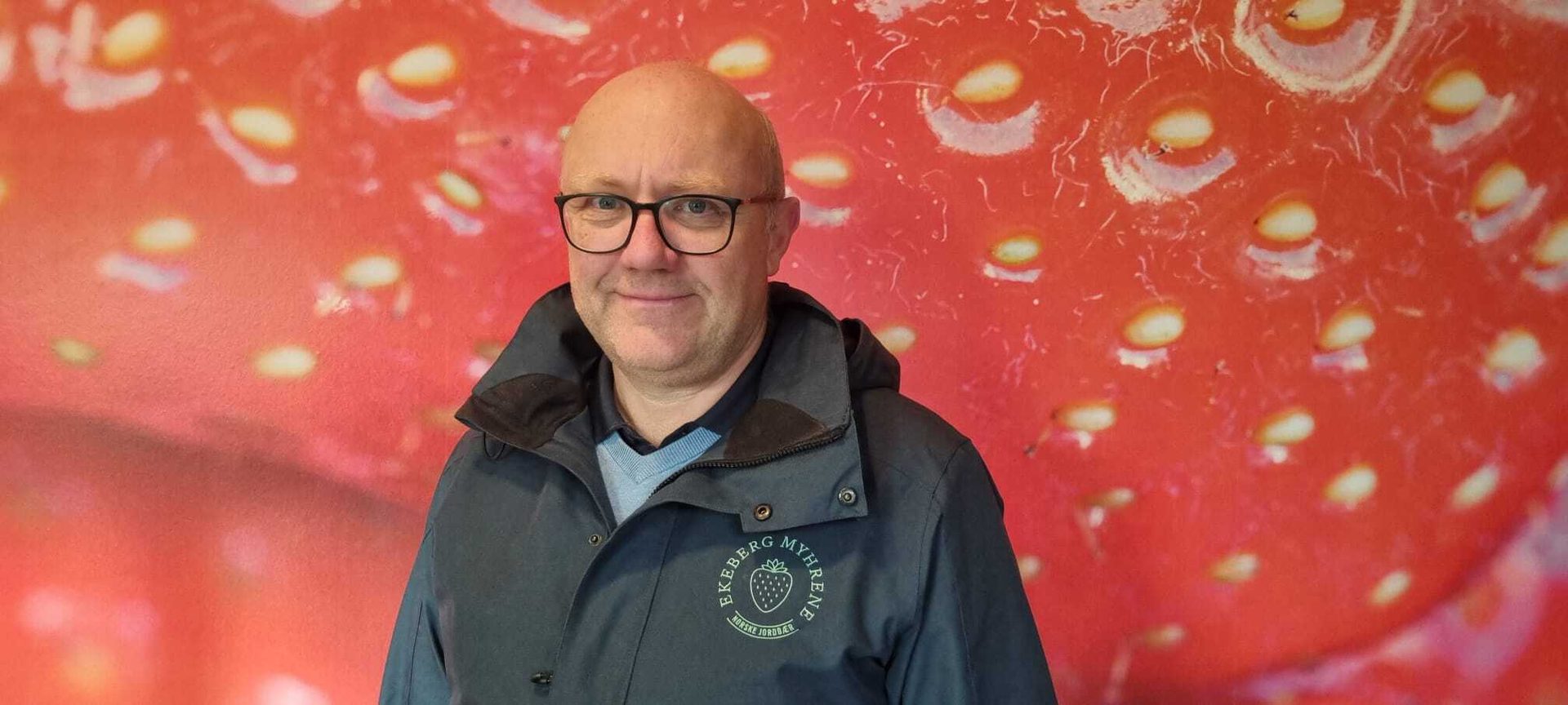 Smiling man in glasses and jacket with strawberry logo against a red, magnified strawberry background.