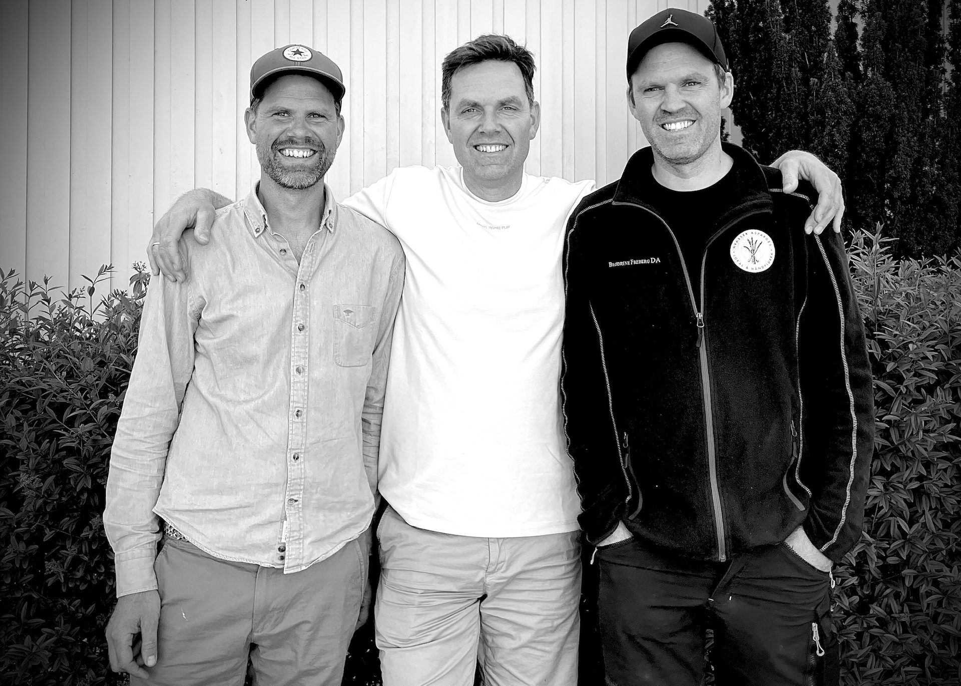 Three adult men, smiling and casually dressed, standing shoulder-to-shoulder with arms around each other.