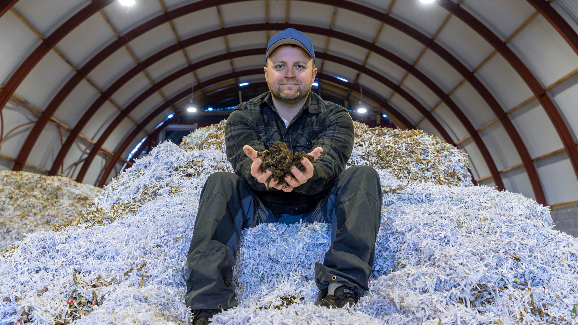Man sits on a large pile of shredded paper, holding soil in his hands, inside an industrial building.