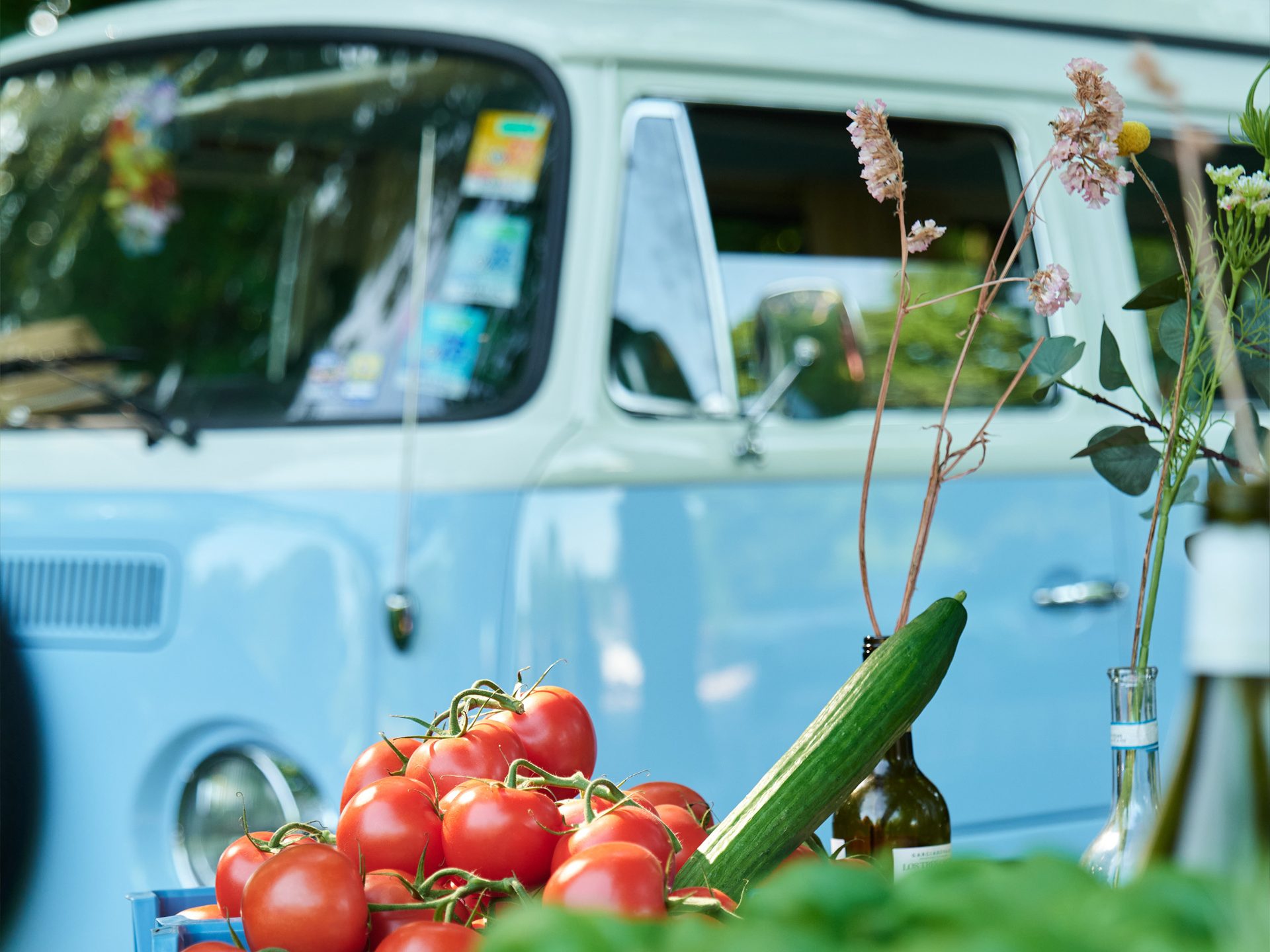 Fresh red tomatoes and a green cucumber in front of a blurred blue and white vintage Volkswagen van.