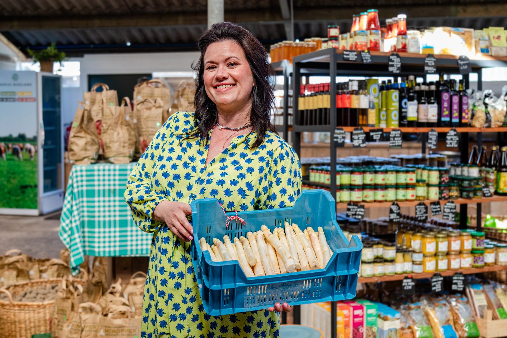 Smiling woman holds a blue crate of white asparagus in a farm shop.