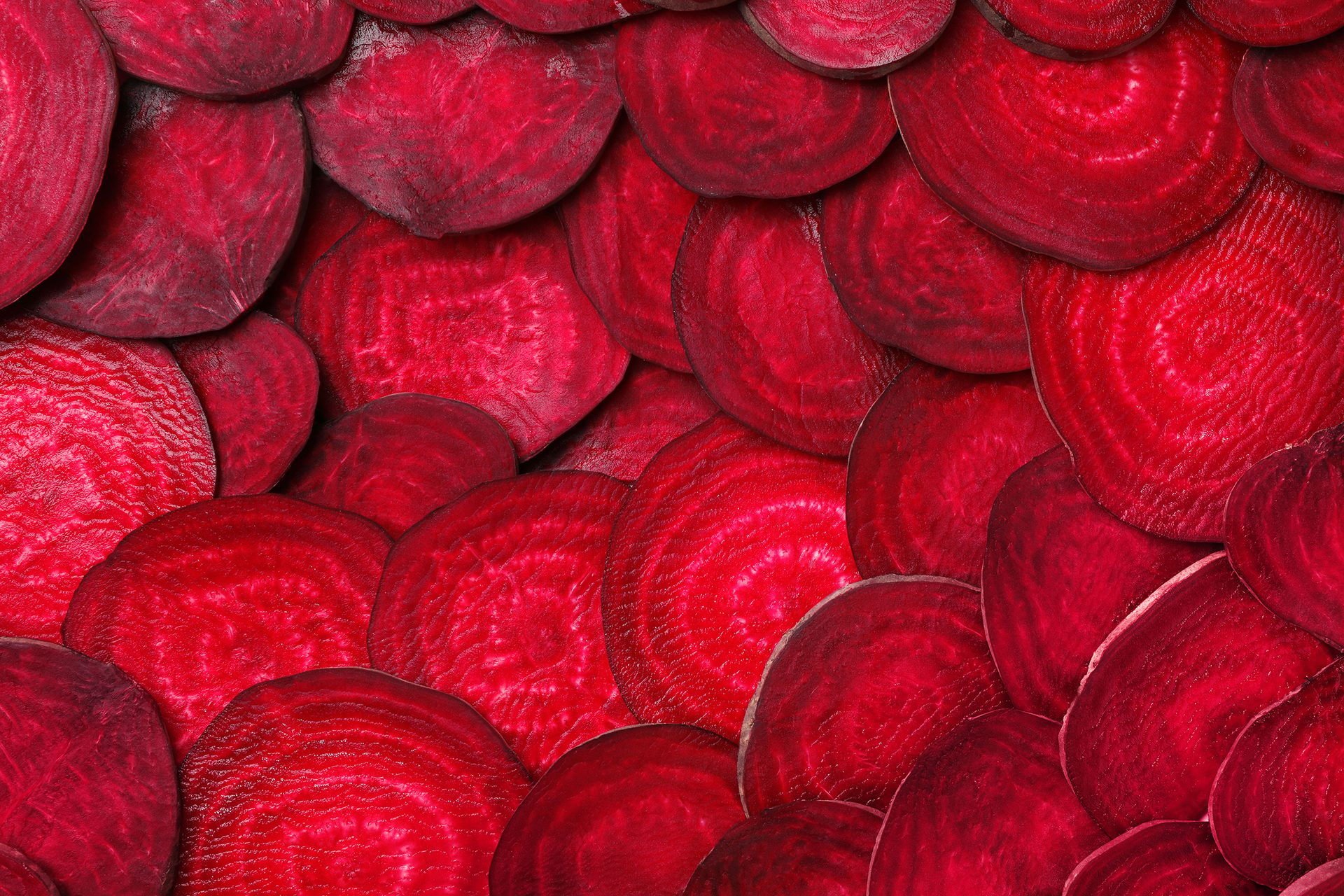Overlapping, thinly sliced raw red beets forming a vibrant, textured background of deep red and magenta.