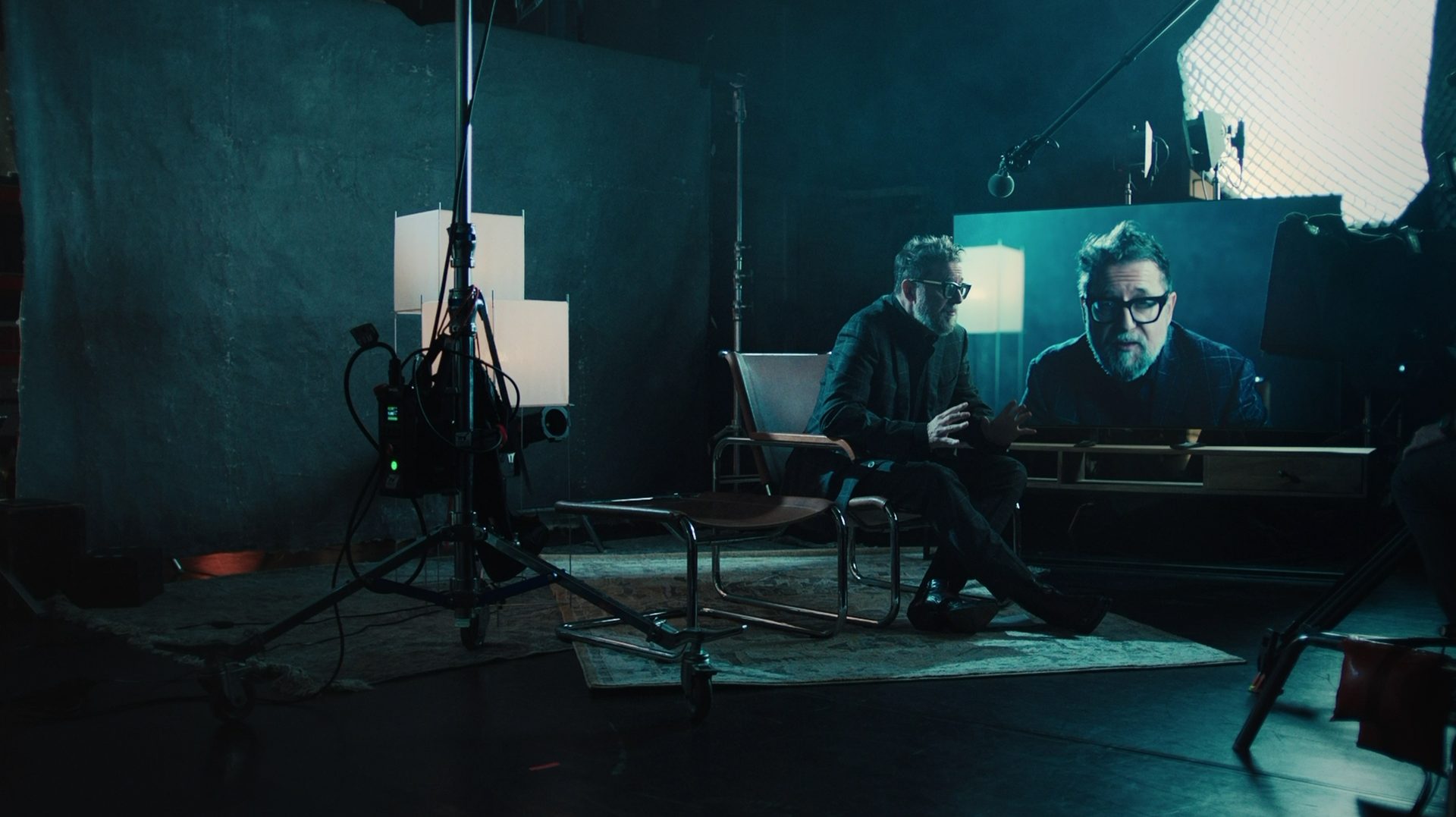 A man sits in a chair on a film set, his image displayed on a monitor nearby.