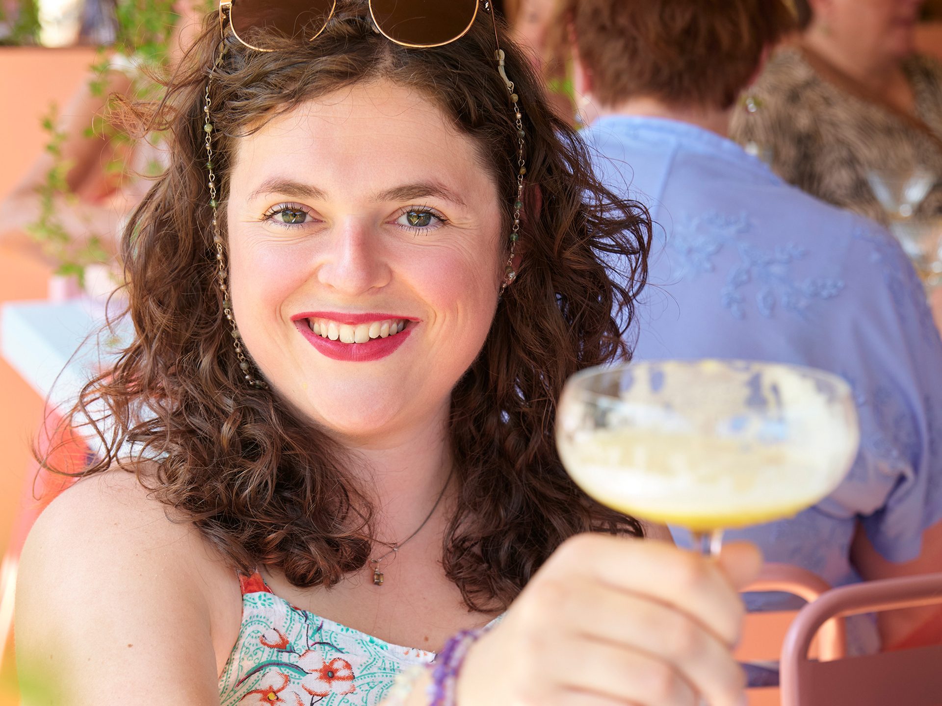 A smiling woman with curly hair and sunglasses on her head holds a cocktail glass outdoors.