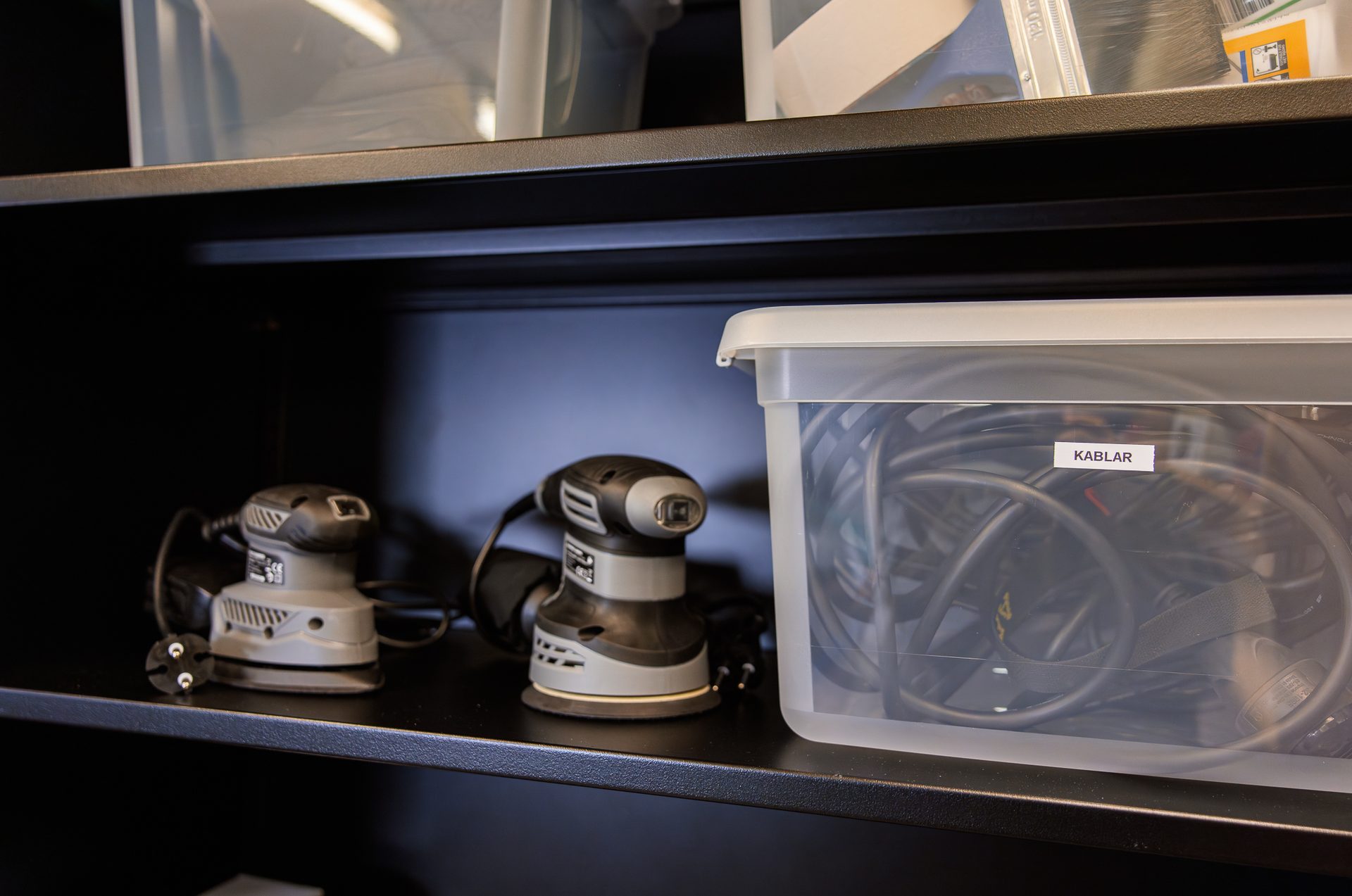 Two power sanders and a clear container of cables labeled "KABLAR" on a dark shelf.
