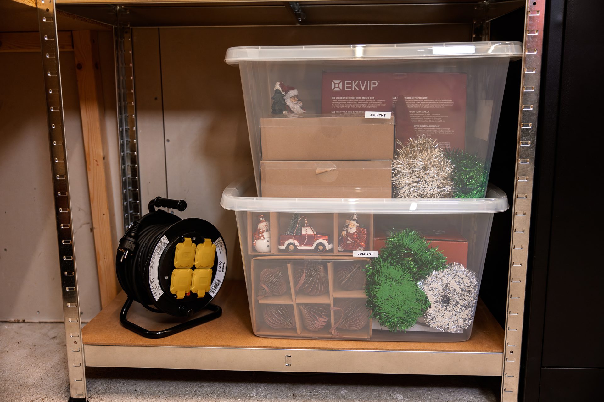 Two clear plastic storage bins filled with Christmas decorations sit on a wooden shelf next to a cable reel.