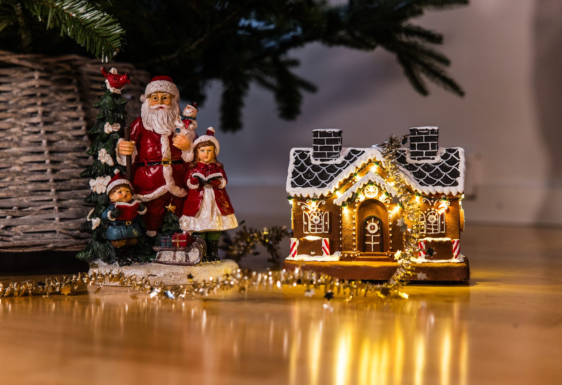 Christmas figurines: Santa with children, a lighted gingerbread house, and sparkling garland on a wooden floor.