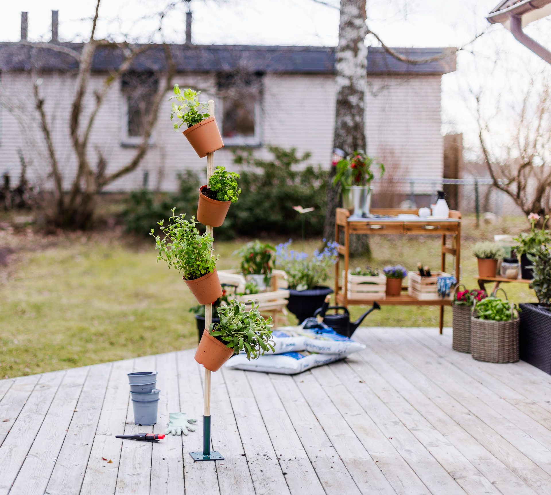 A vertical planter with potted herbs on a wooden deck, with a potting bench and garden in the background.