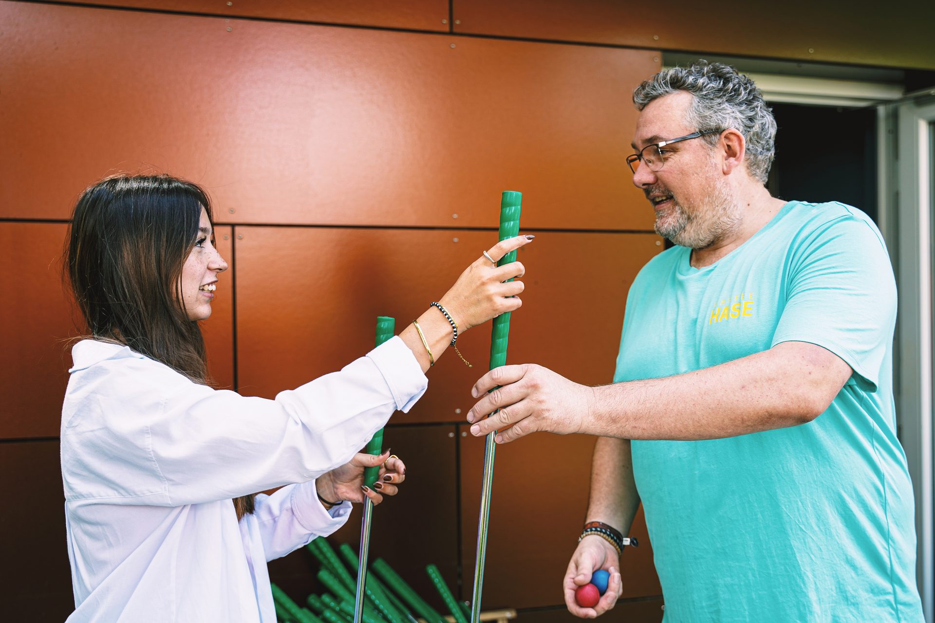 Two smiling people hold green putters; a man also holds red and blue balls.