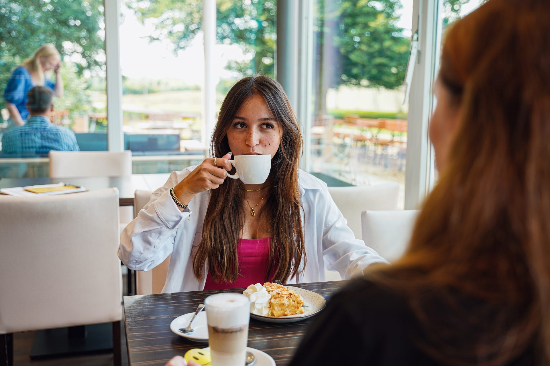 Young woman sips from a white cup at a cafe, with a dessert in front and another person.