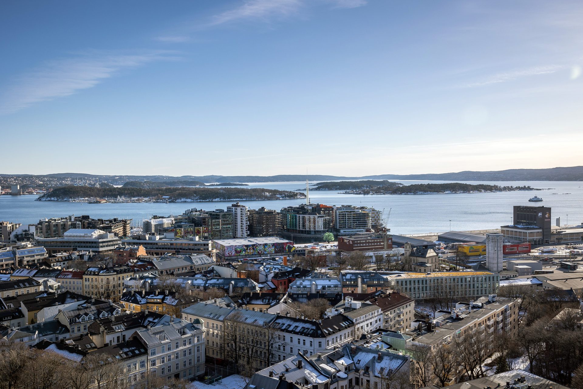 Panoramic view of a snowy city by a fjord with islands and hills under a clear blue sky.