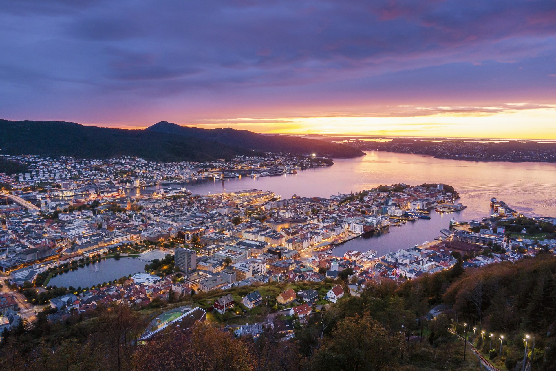 Elevated view of a city at dusk, lights on, by water and mountains, under a vibrant sunset.
