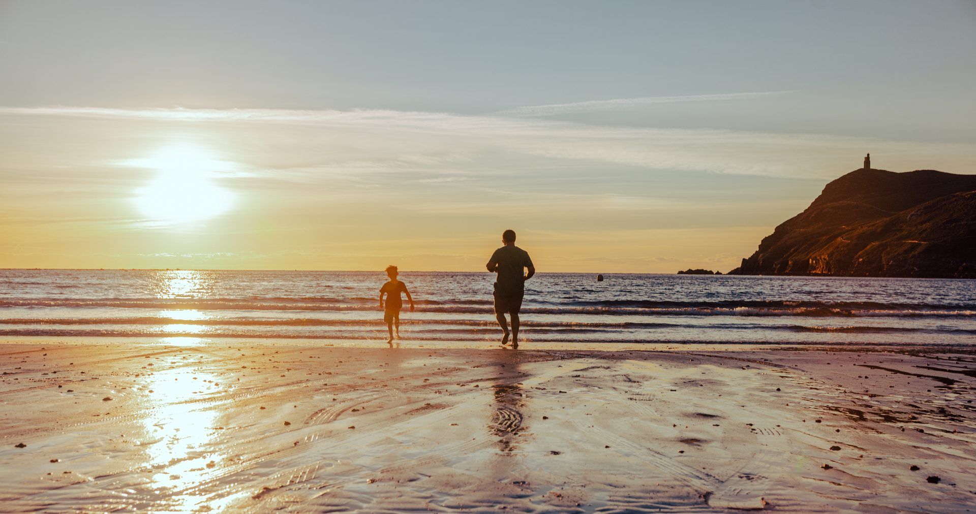 Silhouettes of a man and a child running on a beach at sunset, with a headland in the distance.