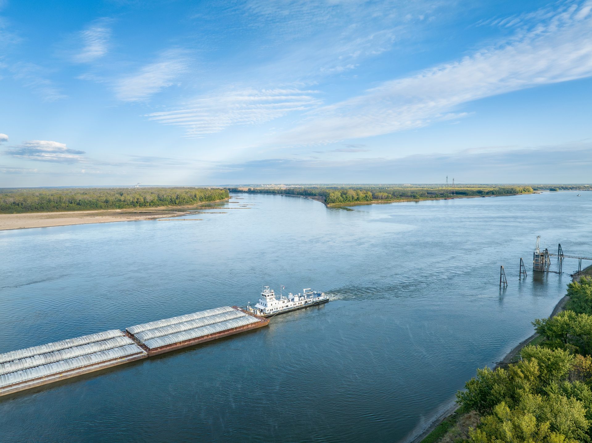 Aerial view of a barge train on a wide river with green banks, under a clear blue sky.