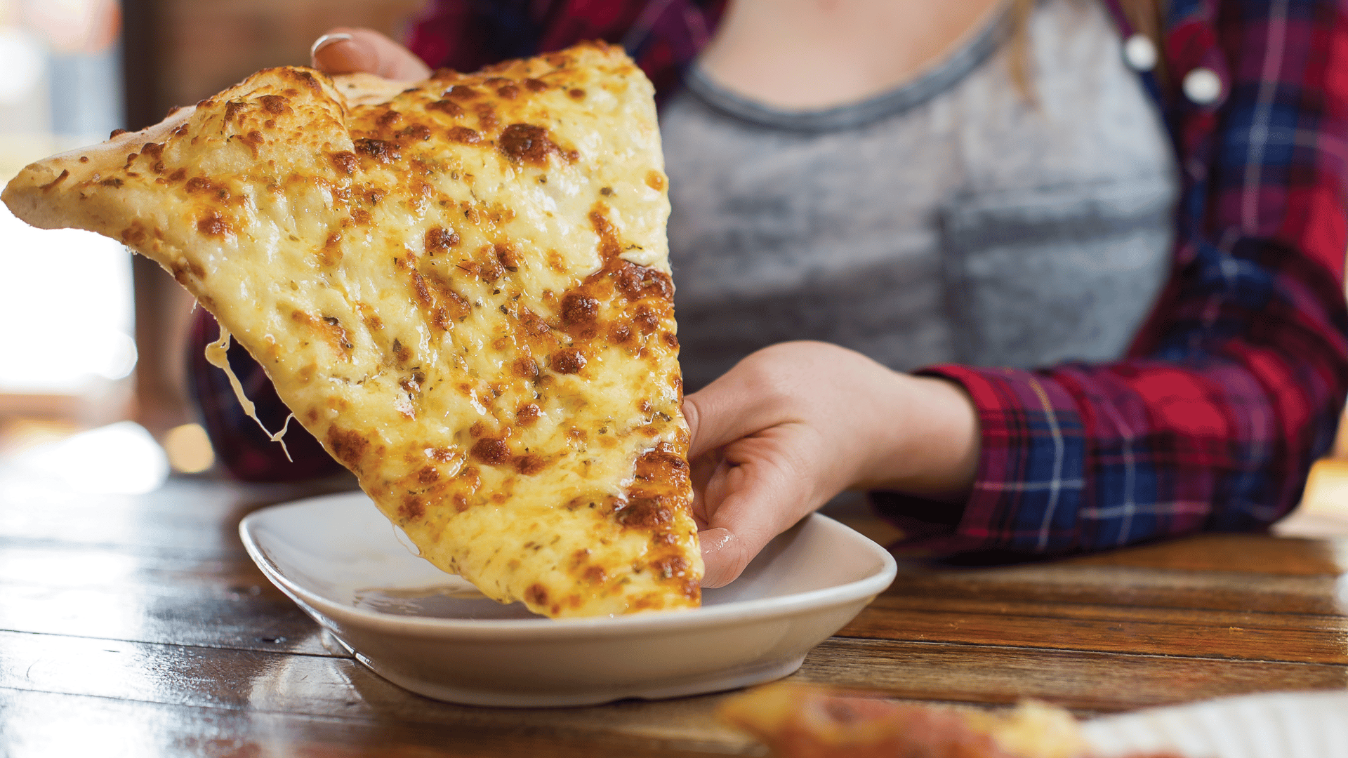 A person in a plaid shirt holding a cheesy pizza slice, with melted cheese stretching.